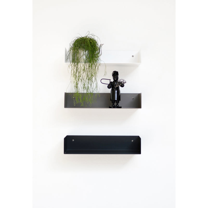 Minimalist wall shelves by Atelier Haussmann create a sleek look: a white steel shelf with a cascading plant, a black powder-coated one showcasing a figurine, and the Poggibonsi shelf remains empty against the white wall.