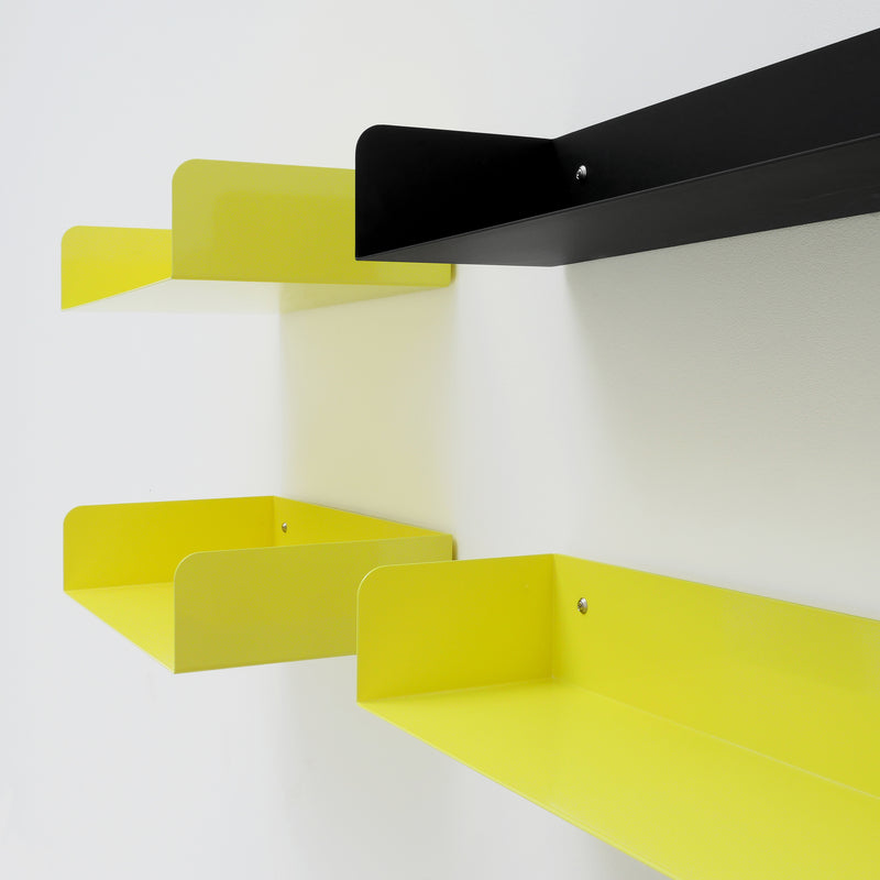 The Poggibonsi Shelf by Atelier Haussmann features four floating shelves mounted on a white wall with a powder-coated finish. Three are yellow, one black, and they are staggered for a minimalist design that embodies vibrant modernity.