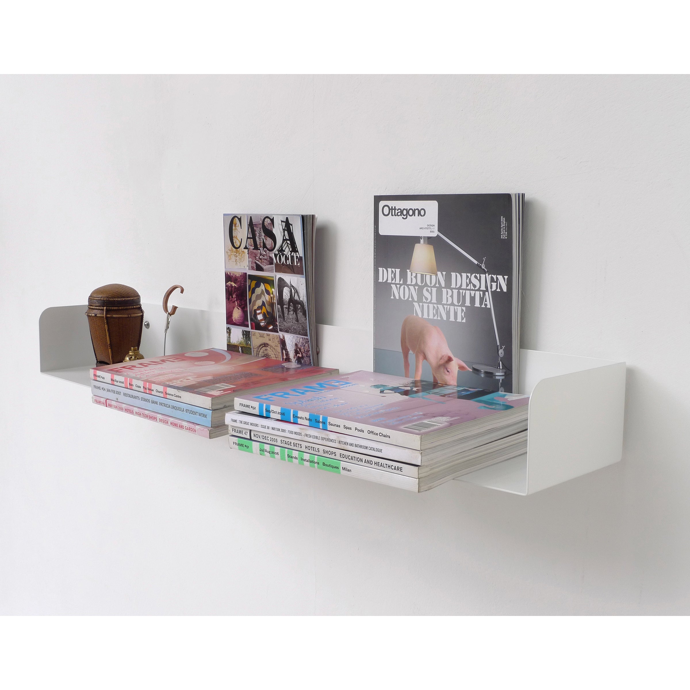 A white, powder-coated Poggibonsi Shelf by Atelier Haussmann displays design magazines stacked upright, showcasing Italian titles. A small wooden container sits on the left. The minimalist and modern wall-mounted shelf highlights the sleek decor.