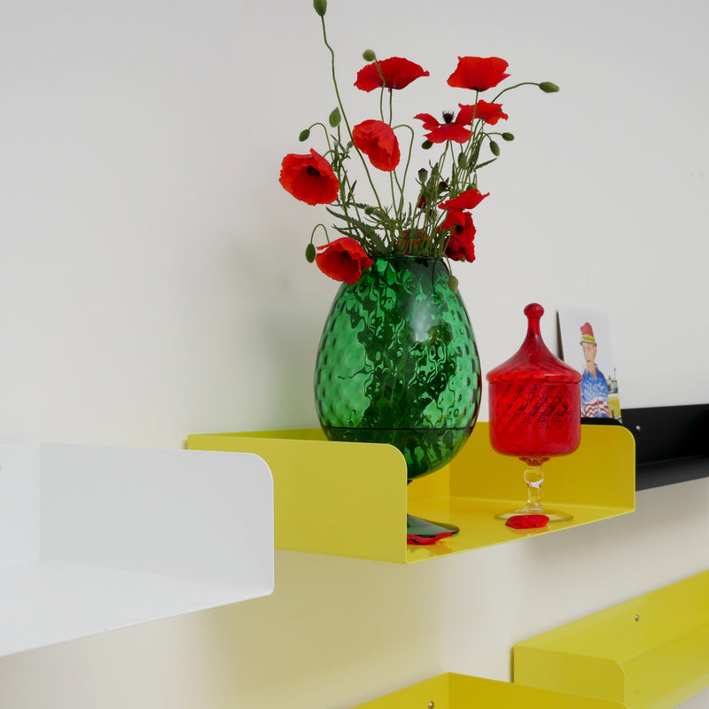 A decorative Poggibonsi Shelf by Atelier Haussmann features a green vase with red poppies, a small red glass container, and a photo frame. The powder-coated yellow shelf is mounted on a white wall among other empty shelves in white, yellow, and black.