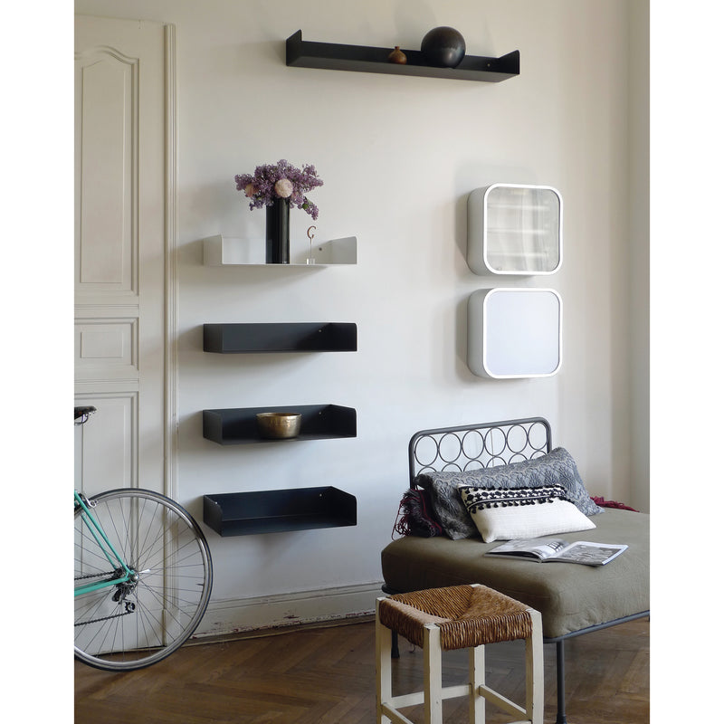 A stylish living room boasts a patterned floor and sleek Atelier Haussmann Poggibonsi Shelf displaying a black vase with purple flowers and a bowl. A nearby beige daybed with cushions and a small woven stool provide comfort, while a bicycle rests against the wall.