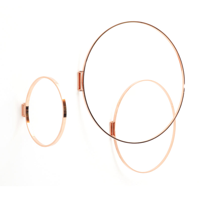 Three overlapping rose gold-finished metal rings by Atelier Haussmann adorn a white wall. These Rings, varying in size, create a modern, minimalist design, resembling an elegant art piece.