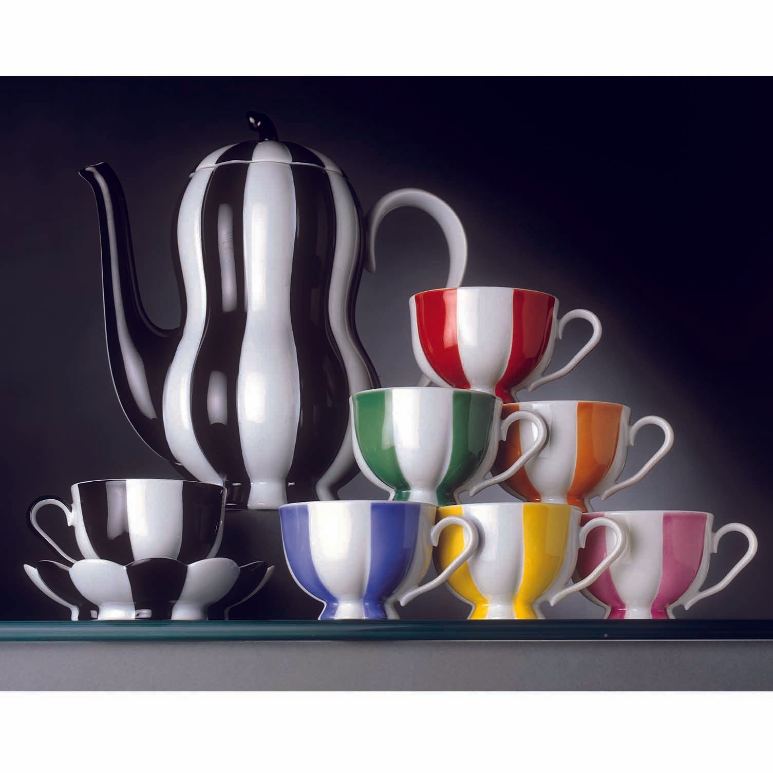 A colorful set of Augartens Melon Mocha Cups, featuring red, green, orange, blue, yellow, and pink two-toned designs with white saucers, is stacked next to a matching black-and-white striped teapot on a glass shelf.