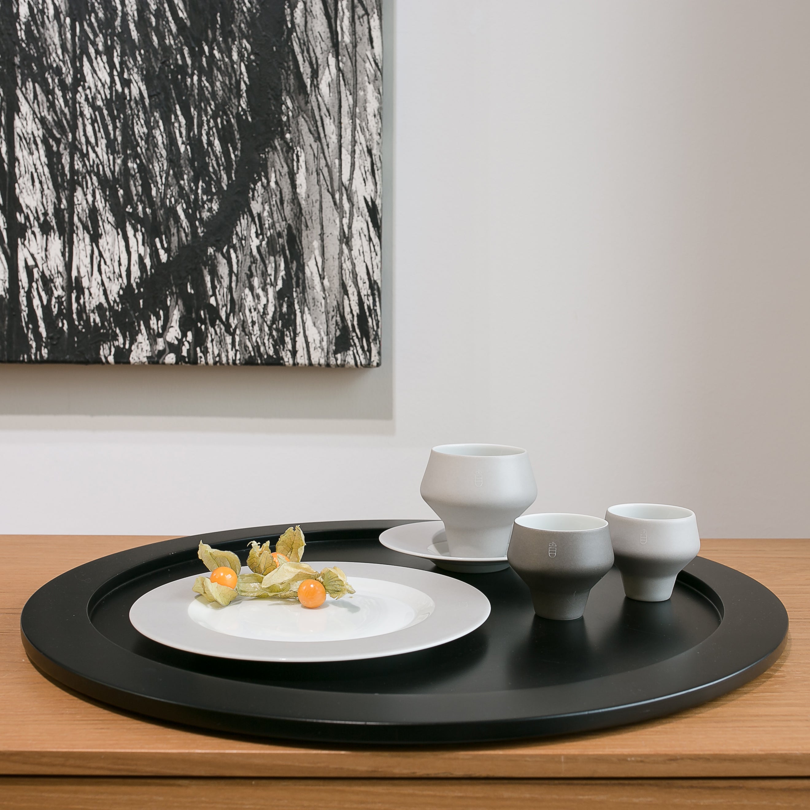 A modern still life features three white ceramic cups from Augartens Shortcut collection on white plates with a black tray. Physalis fruits are on the foreground plate. A monochrome abstract painting is in the background, all elegantly arranged on a wooden surface.