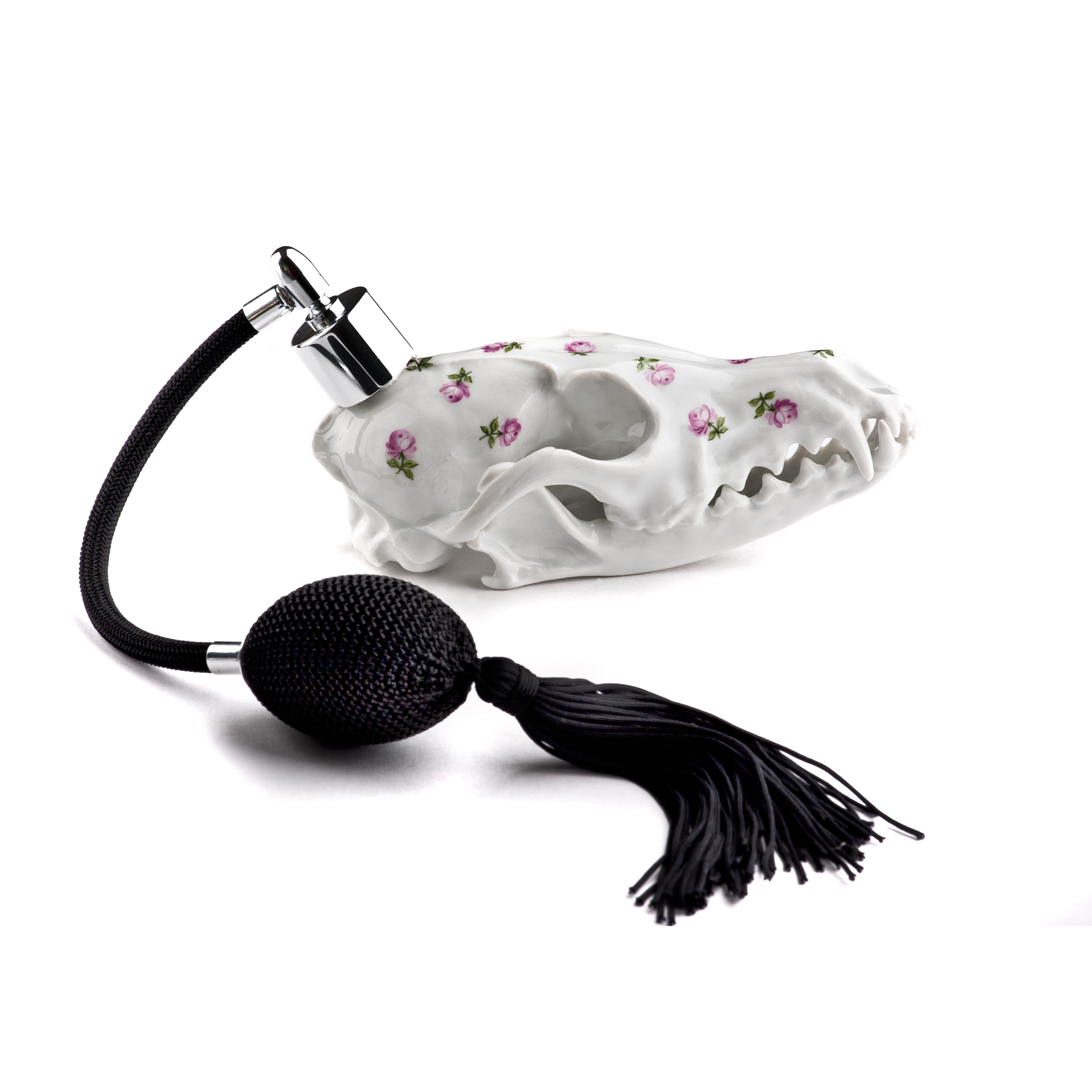 The Vulpini Flacon: Rose by Augarten features an animal skull-inspired porcelain design with pink flowers. Its Vienna Porcelain Manufactory origins shine through in its hand-painted elegance, enhanced by a black atomizer with tassel on a white background.