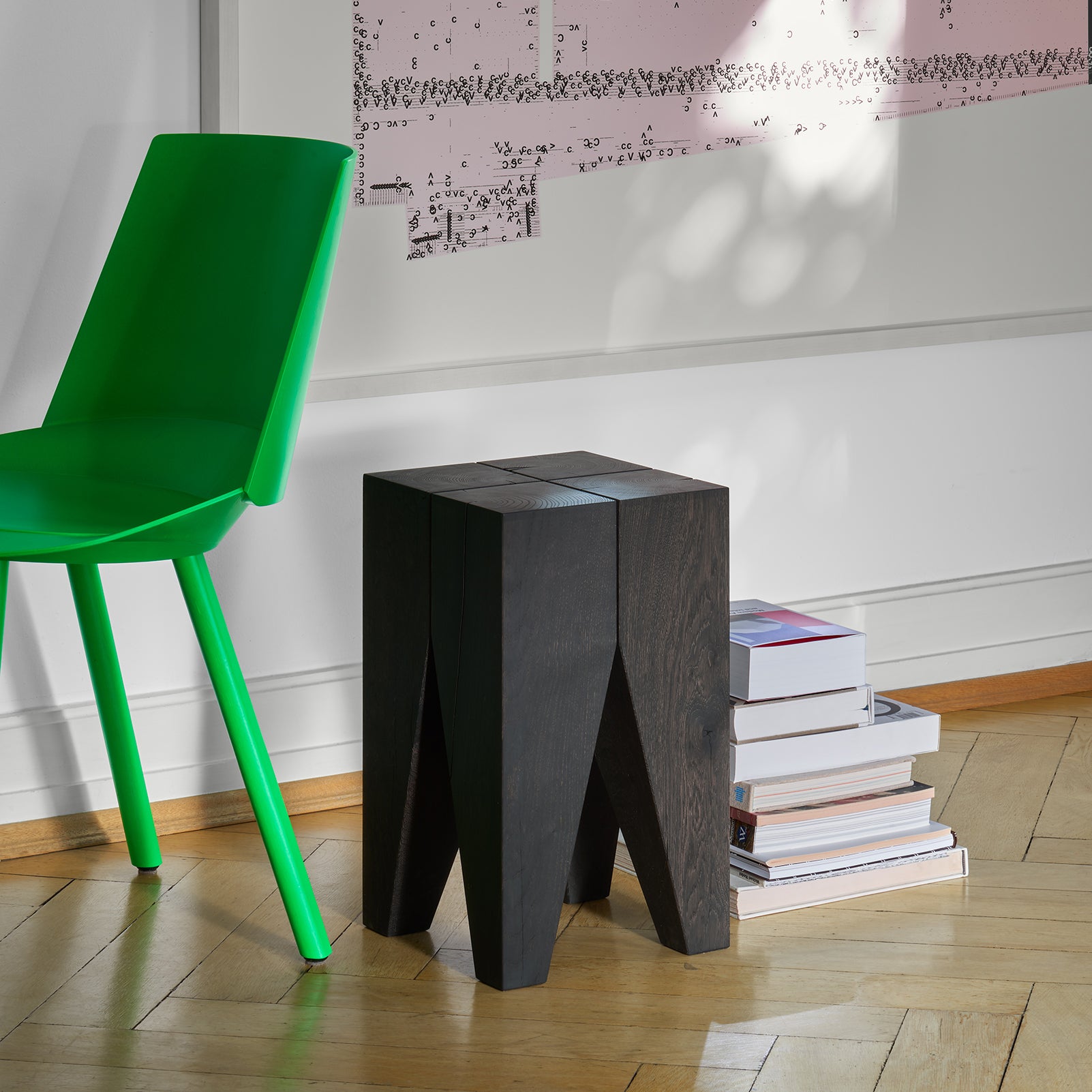 A modern green chair on a wooden floor is paired with E15s sleek, solid wood ST04 Backenzahn Black Oak stool. A stack of books is nearby, while abstract wall art decorates the light wall in the background. Light filters through, casting intricate shadows and enhancing this classic design scene.