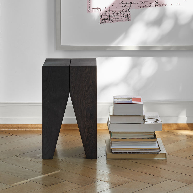The minimalist interior features E15s ST04 Backenzahn Black Oak stools beside a stack of books on a wooden floor. A large artwork with mathematical equations adorns the white wall, partially lit by dappled sunlight, creating a sleek and timeless ambiance.