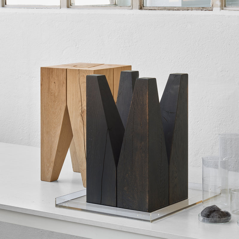 Two solid wood stools sit on a white surface. The front classic features a dark V-shape design, while the E15 ST04 Backenzahn Black Oak stool is behind it in light wood. Both are in a minimalistic space with glass jars and a white wall backdrop.