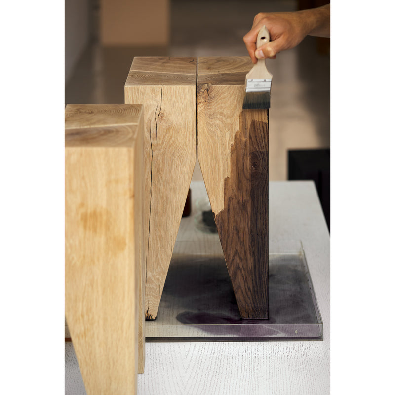 A person uses a brush to apply a dark stain to the E15 ST04 Backenzahn Black Oak stool, subtly transforming its light wood color. Positioned on a white surface, its staining process is accentuated by the softly blurred background, highlighting this design classic.