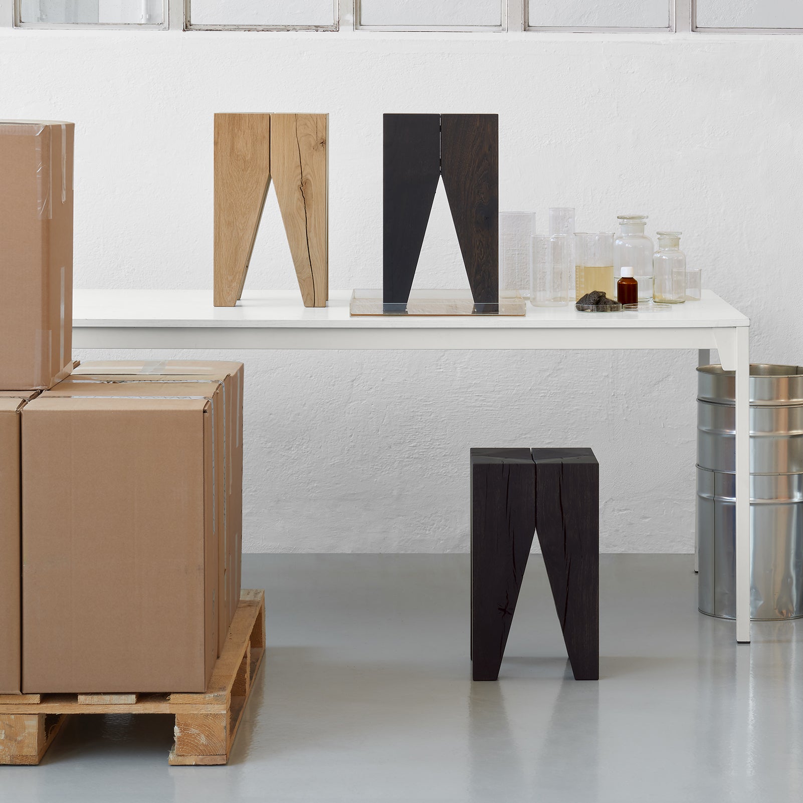 In a white room, an E15 table showcases Black Oak ST04 Backenzahn stools beside variants in natural and dark brown finishes. Cardboard boxes sit on a pallet upfront, with glass containers and metal drums providing an industrial backdrop.