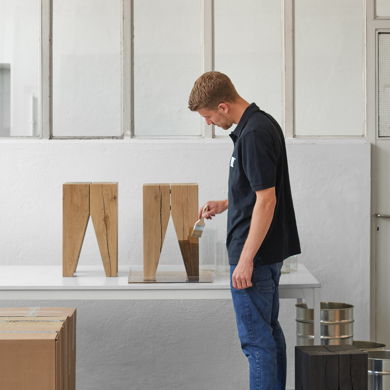Wearing a black shirt and blue jeans, a man paints a small wooden table named ST04 Backenzahn Black Oak by E15, reflecting classic elegance. Nearby, two similar tables and paint tins sit on a white surface, with large windows and a cardboard box completing the creative scene.