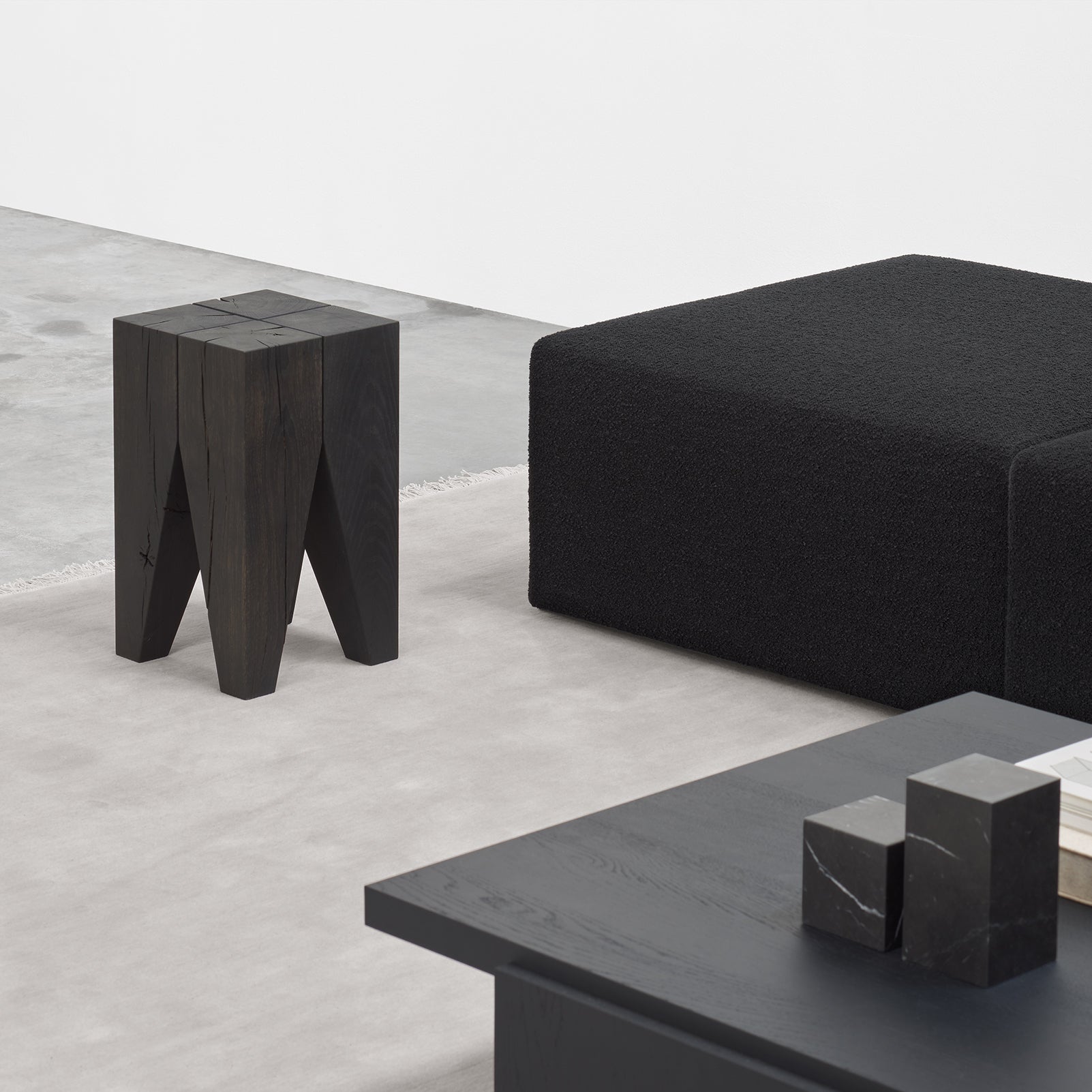 A minimalist living space includes a modern black cubic sofa, an E15 ST04 Backenzahn Black Oak stool with a split design, and a sleek black coffee table with geometric blocks. The floor is light grey concrete.