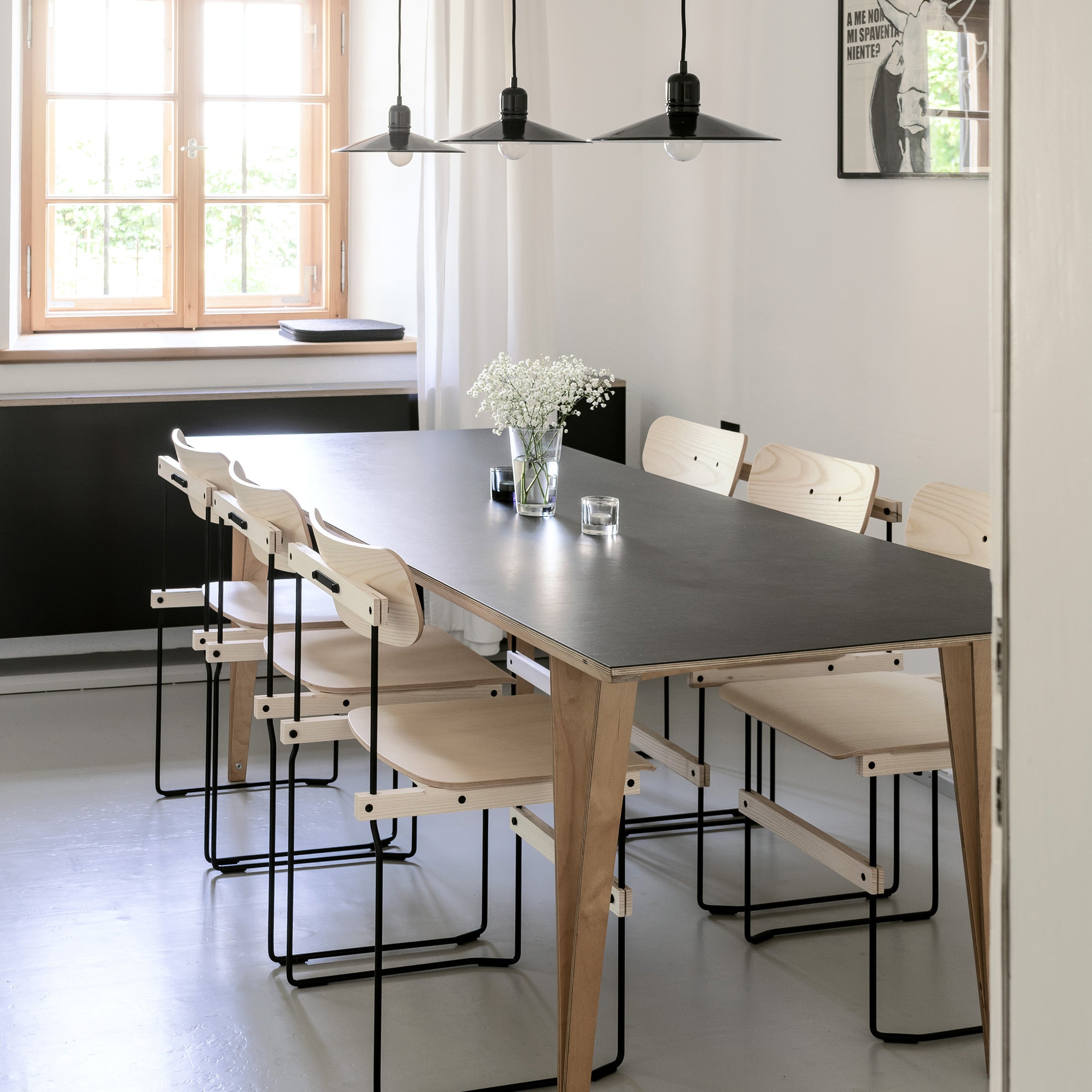 A modern dining room highlights the sleek design trend with a Spanoto table from Moormann, featuring a black surface and light wooden legs. Matching wooden chairs with black metal frames encircle it, while minimalist pendant lights hang overhead. A window and framed artwork complete the scene.