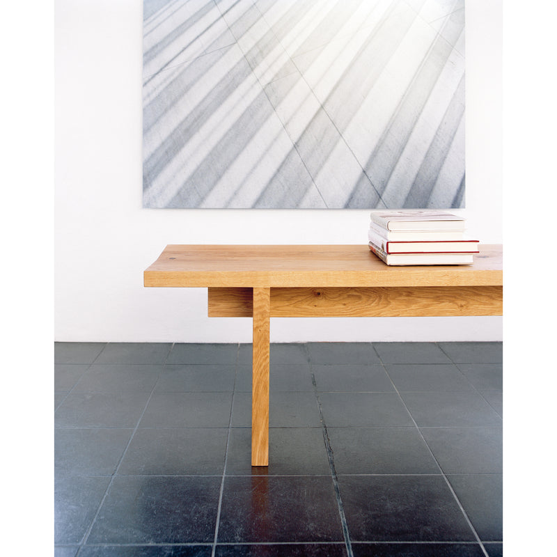 A minimalist interior includes a wooden table with books, accompanied by the E15 BE02 TARO bench. The dark tiled floor enhances the large abstract grayscale artwork on the white wall behind it, reflecting Philipp Mainzers refined aesthetic.
