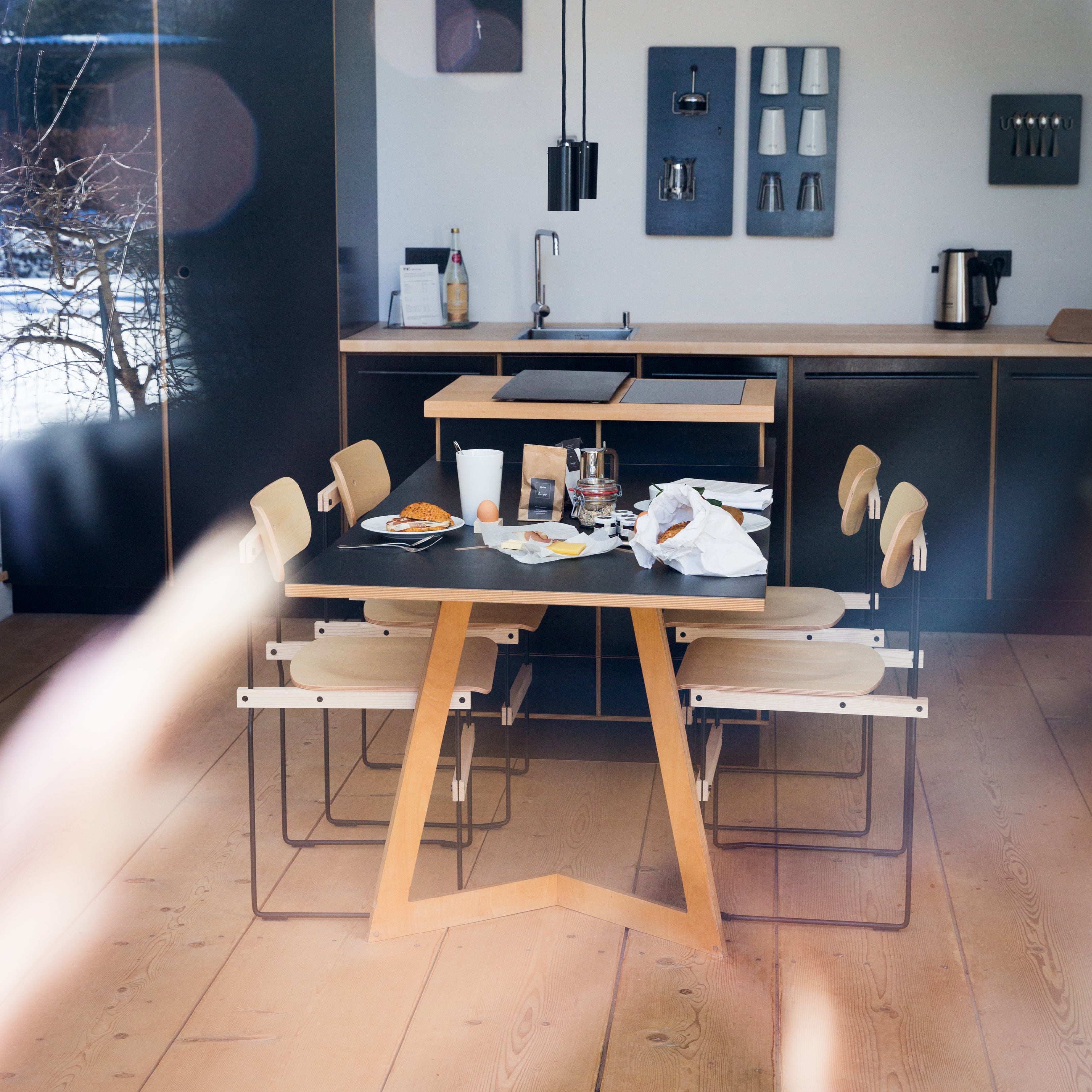 A modern kitchen showcases a Bruto wooden table set for breakfast with molded plywood chairs. Plates, cups, and breakfast items adorn the table. In the background are a sink, sleek black cabinets, and a coffee maker bathed in sunlight from the window. Brand: Moormann.