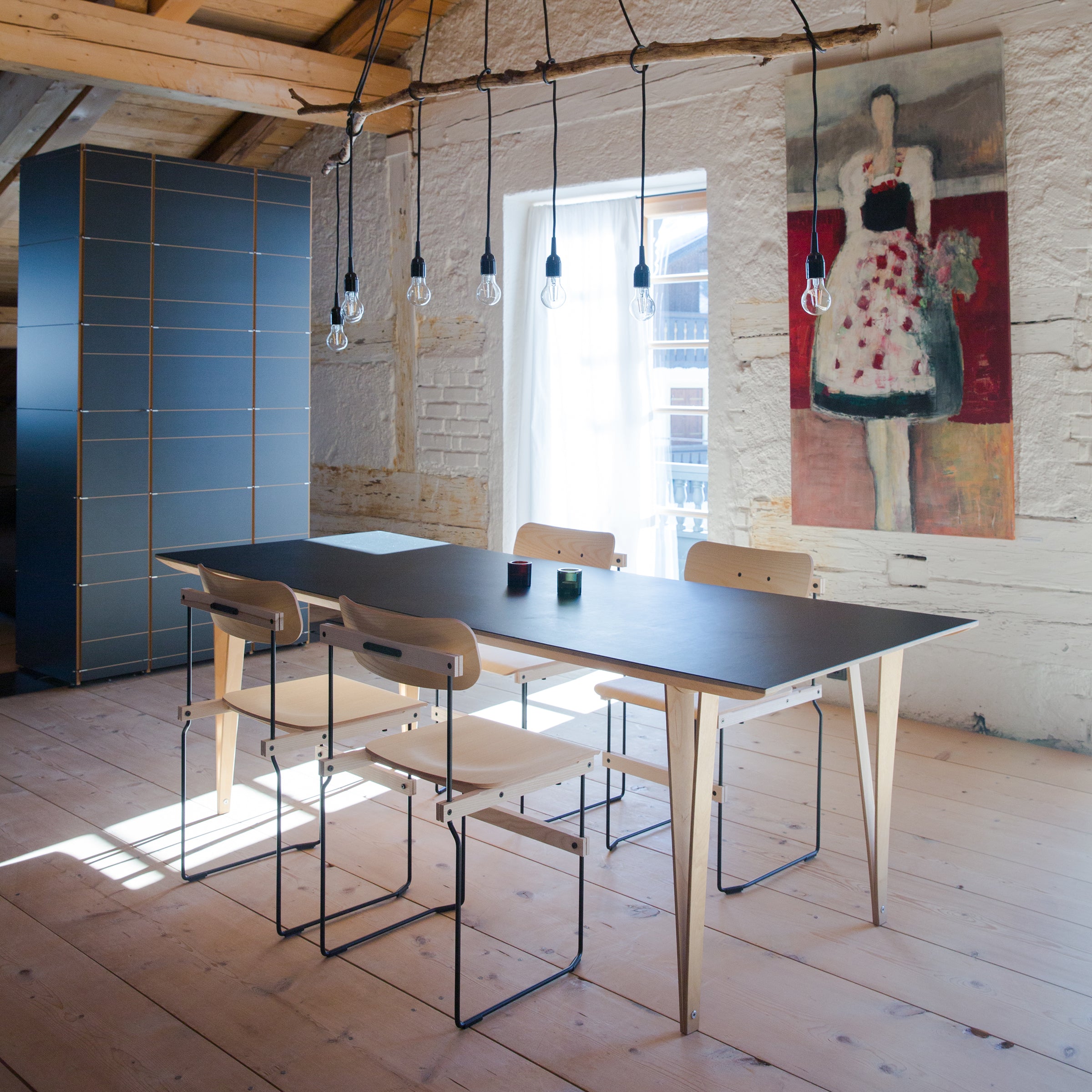 A contemporary dining space includes Moormanns Spanoto, a birch plywood wooden table, surrounded by four matching chairs on a similar floor. Exposed brick walls and a large painting add charm, with pendant lights above the table and sunlight streaming through the window.