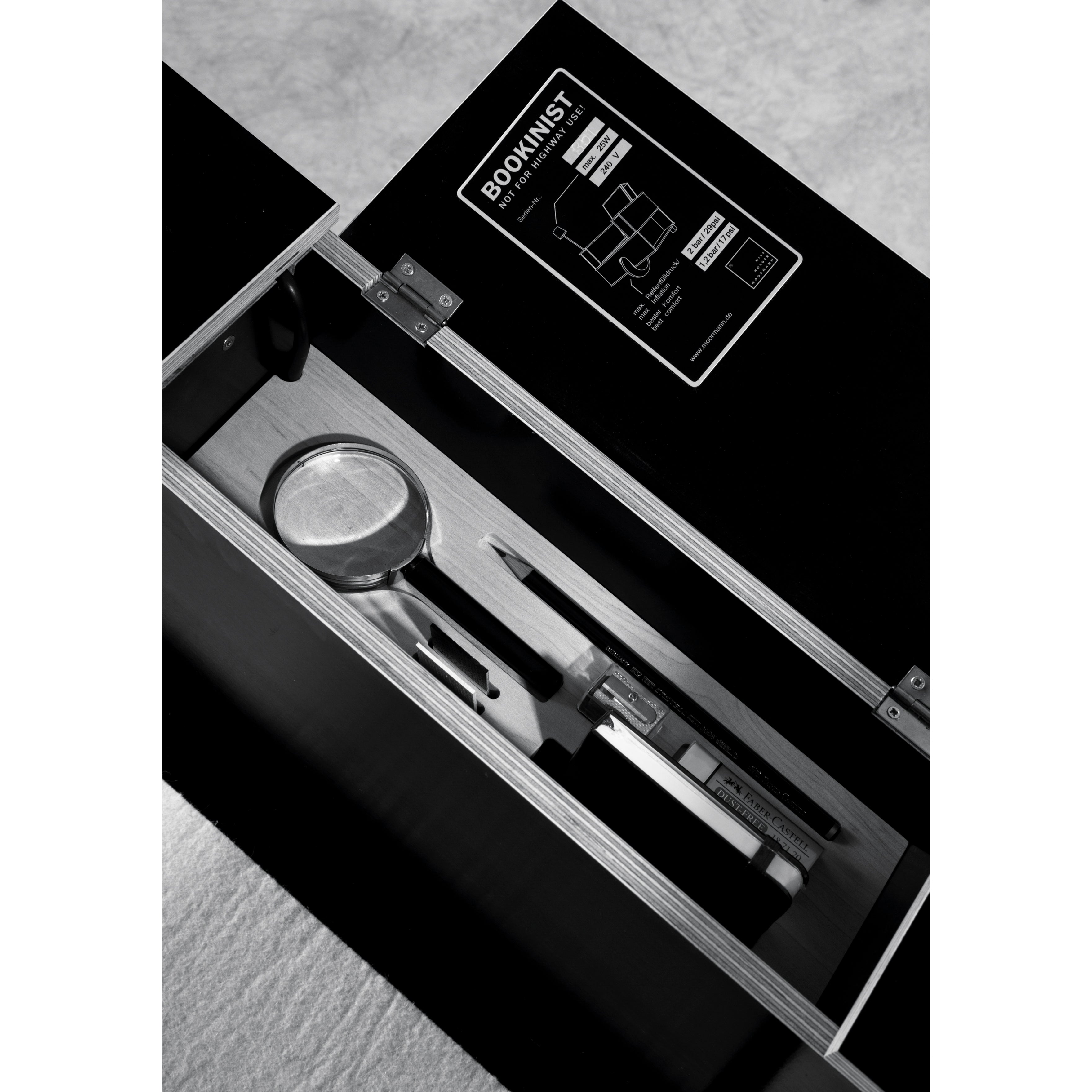 A black and white photo showcases an open box labeled Bookinist by Moormann, containing a magnifying glass, pen, and smartphone. Inside the lid is a diagram. The minimalist setting features a textured surface beneath, suggesting its use with a movable chair and smart book storage options.