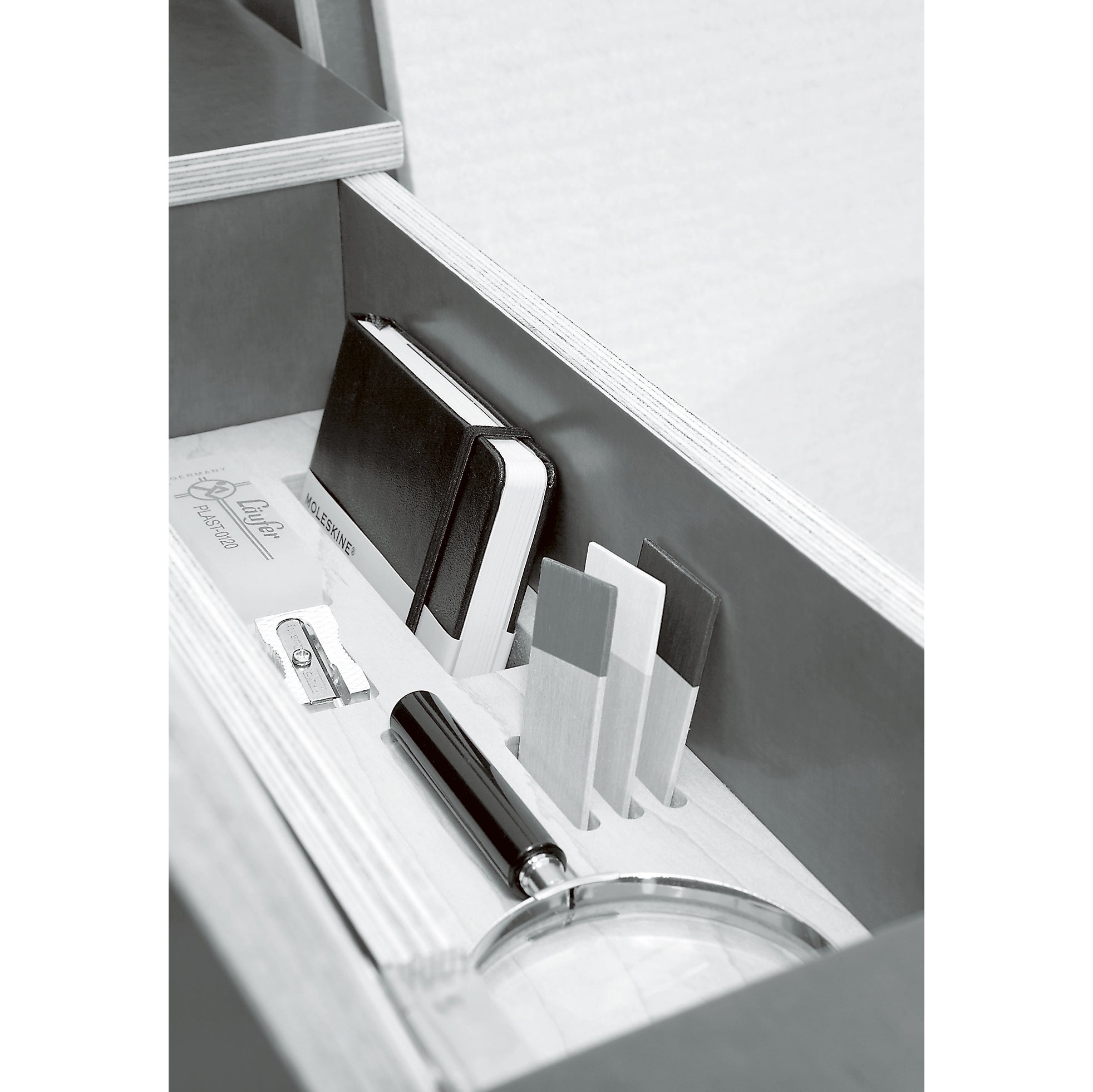 The Moormann Bookinist features a drawer with a notebook, three wooden cards, a pencil sharpener, and a magnifying glass under its sleek white surface. A comfortable reading chair nearby invites exploration.