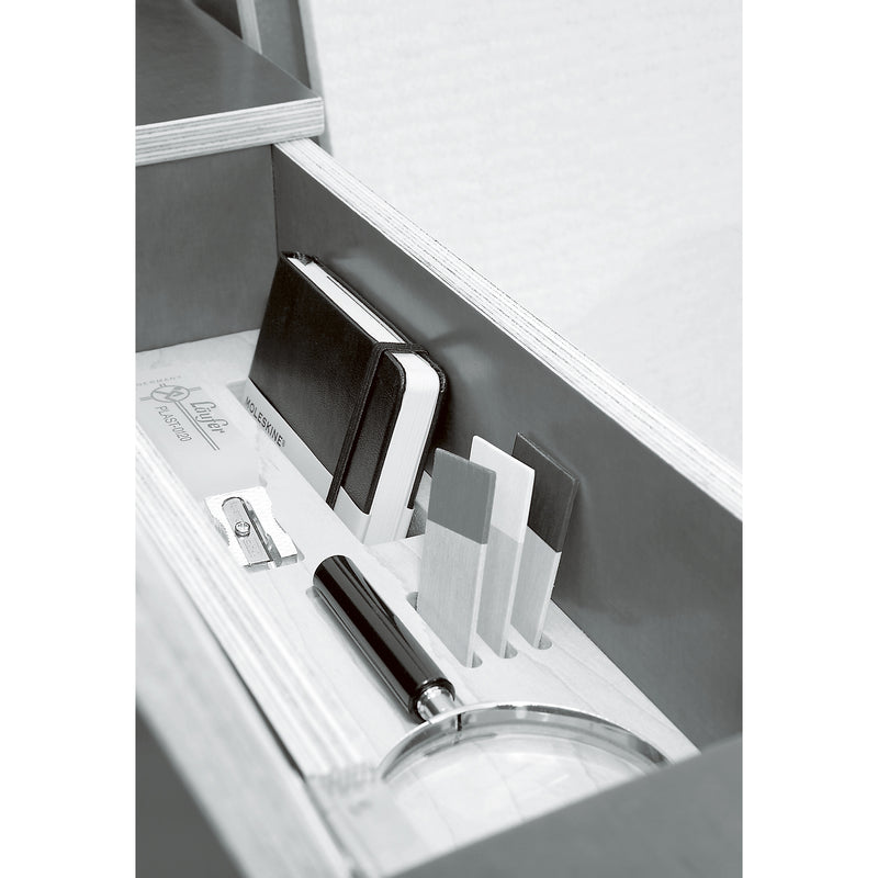 The Moormann Bookinist features a drawer with a notebook, three wooden cards, a pencil sharpener, and a magnifying glass under its sleek white surface. A comfortable reading chair nearby invites exploration.