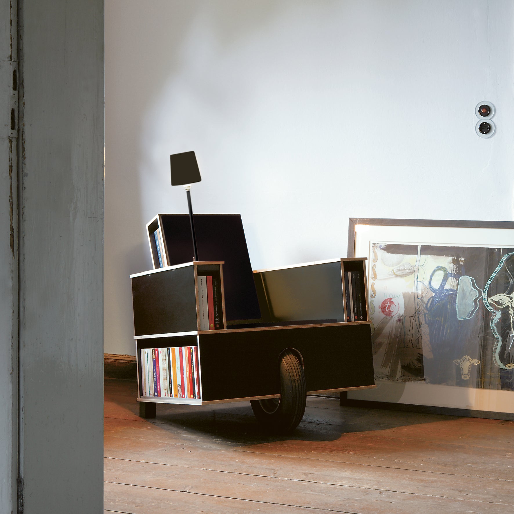 The Bookinist by Moormann is a sleek black reading chair featuring built-in book storage on both sides, a base wheel, and an attached lamp. This mobile chair complements wooden floors with a nearby painting leaning against the white wall.