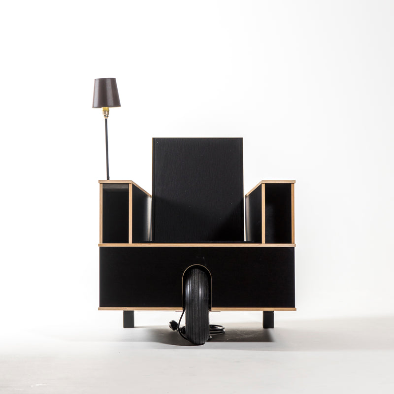 The Bookinist by Moormann is a sleek, modern black reading chair with open side book storage and an attached lamp on the left. It has a wheel at the base for versatility and easy movement, all set against a plain white background.