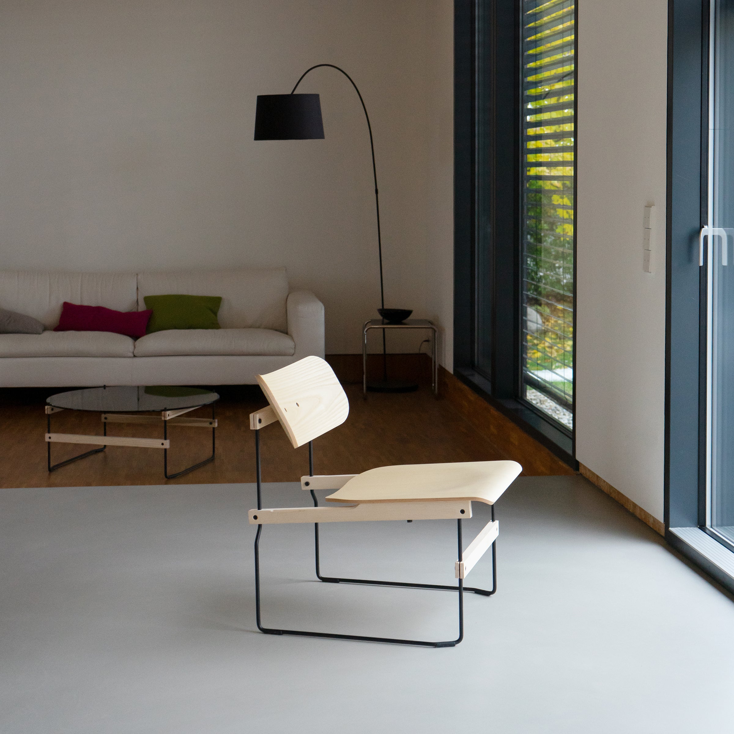 A minimalist living room showcases Moormanns Brutissimo chair with a sleek black metal frame and light molded wood. A white sofa with colorful cushions, a round glass coffee table, and a tall black floor lamp sit near large windows in the background.