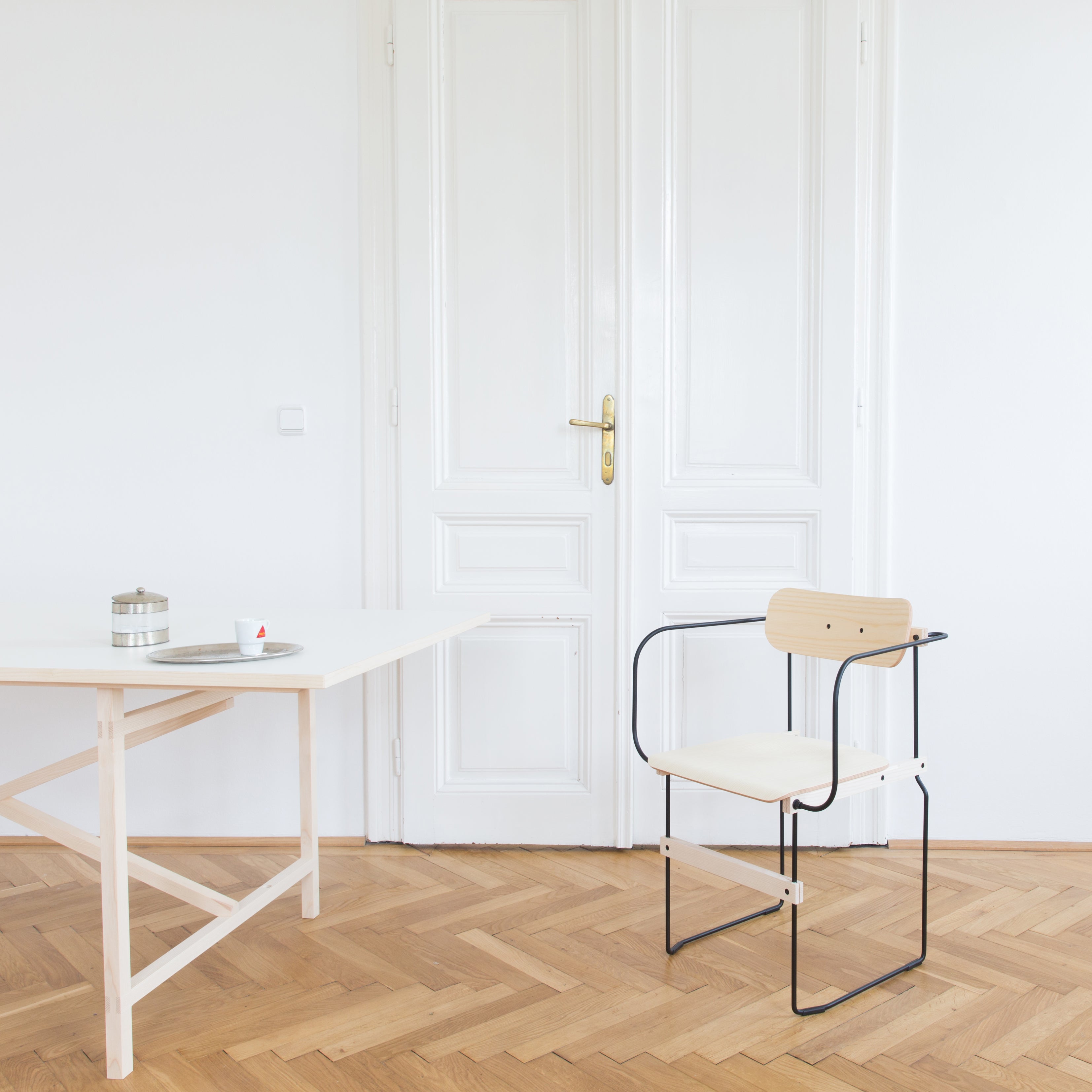 A minimalist room includes a white table with molded wood surfaces, complemented by Moormanns Bruto Armchair on the wooden herringbone floor. A small tray holds a metal container and cup, while the flexible design is framed by a white paneled door and walls in the background.