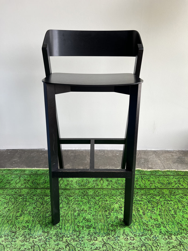 A tall, black Barstool Merano by TON sits on a bright green patterned rug against a plain white wall, highlighting its elegant design crafted from solid wood.