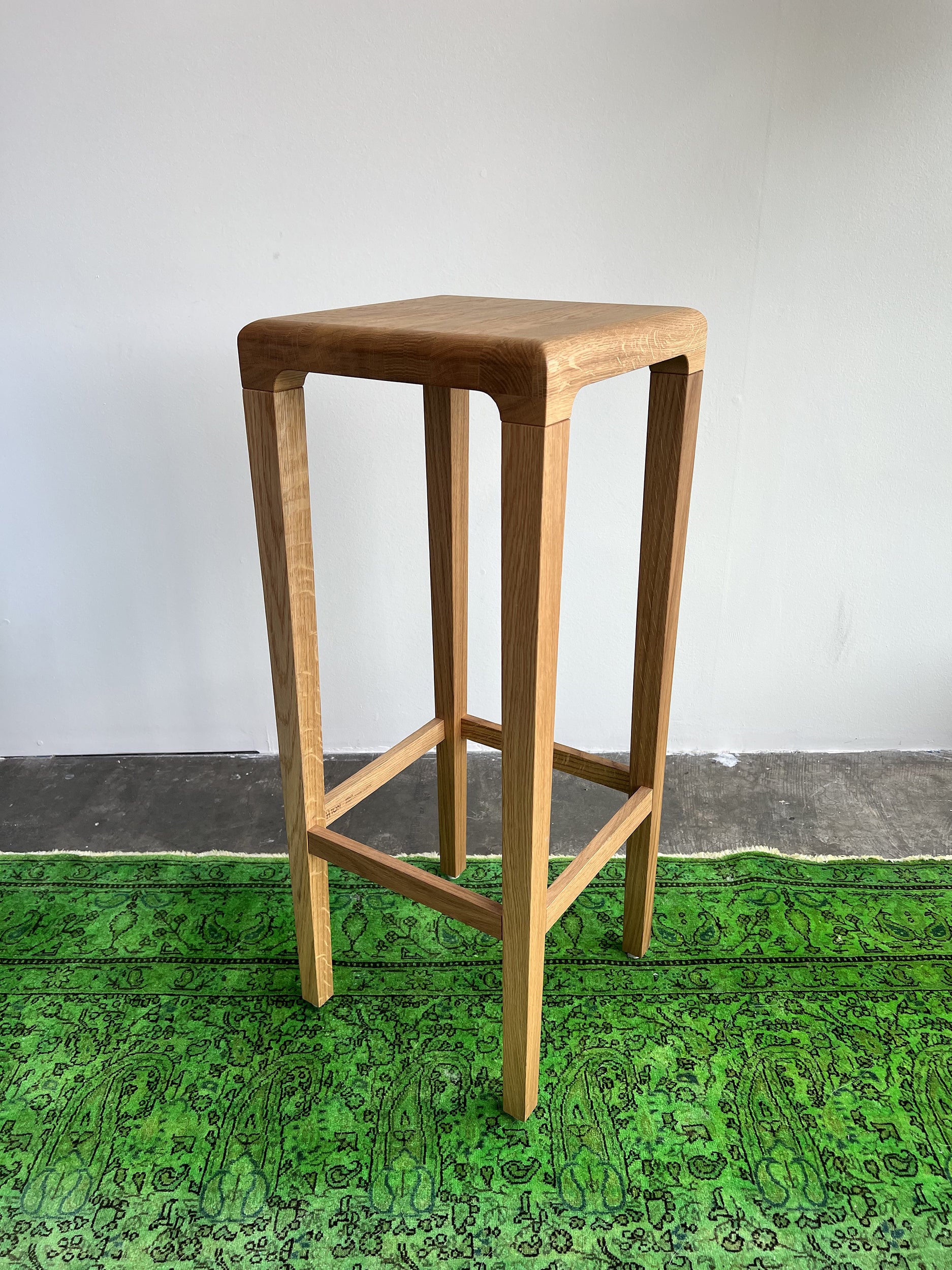 The Barstool Rioja from TON, featuring a wooden square seat with four legs and supporting crossbars, sits atop a green patterned rug. Crafted from oak, it offers a simple modern design against a plain white wall and concrete floor backdrop.