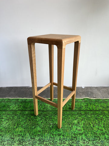The Barstool Rioja from TON, featuring a wooden square seat with four legs and supporting crossbars, sits atop a green patterned rug. Crafted from oak, it offers a simple modern design against a plain white wall and concrete floor backdrop.