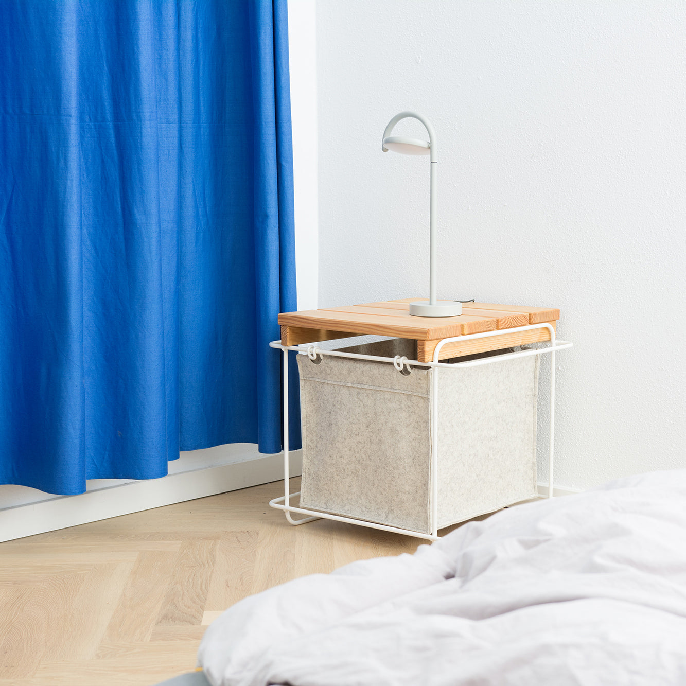 By the bed stands a Grit Indoor nightstand by Bartmann Berlin, featuring a wooden top and white wool felt basket. A minimalistic white lamp sits on it. Nearby, a bright blue curtain adds color against light wood flooring.