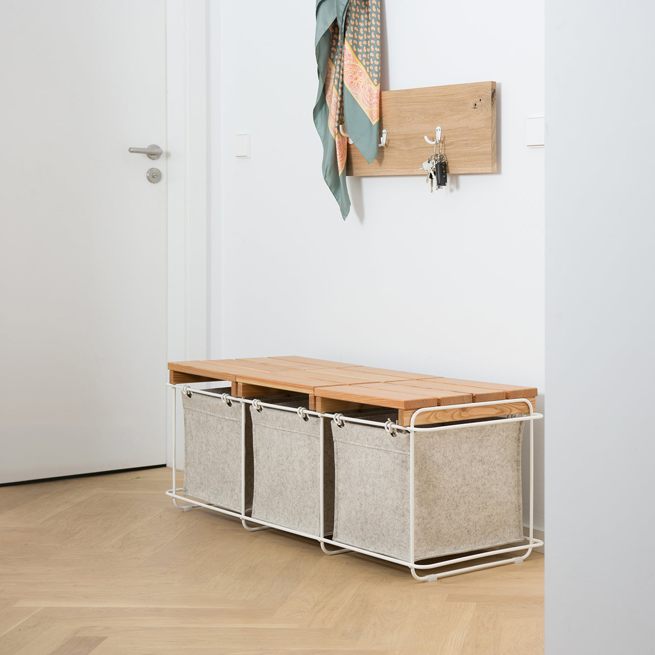 A minimalist hallway features the Grit Stool Indoor by Bartmann Berlin, made of wood and metal. Three beige fabric storage bins are underneath. Above, a wood wall hook holds a scarf and keys. The light wood floor enhances the modern aesthetic with stainless steel accents adding sleekness.