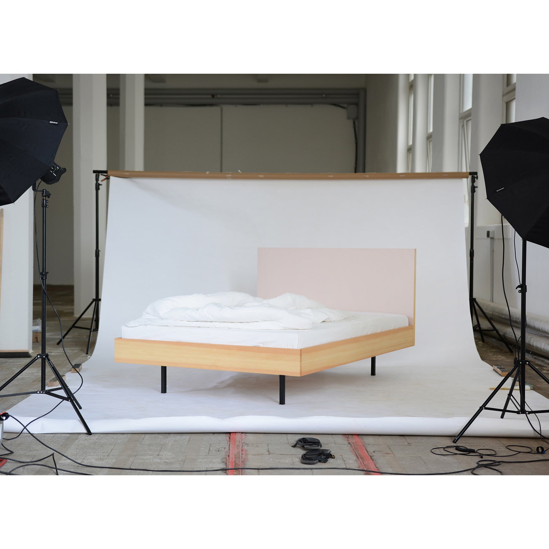 A Bartmann Berlin Unidorm Bed Powder with light wood and white bedding is set up in a photography studio, flanked by two large studio lights on black steel stands against a large white backdrop.