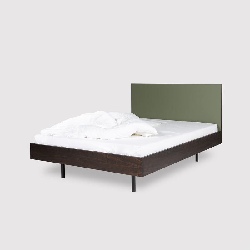 The Unidorm Bed Olive by Bartmann Berlin is a minimalist double bed featuring a dark smoked oak frame, olive green headboard, and black slim legs. It boasts crisp white bedding against a light grey backdrop for a modern design aesthetic.