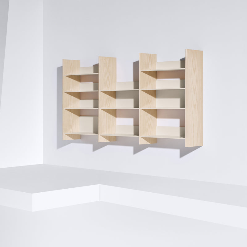 The Graph by Bartmann Berlin is a small, modern wall-mounted bookshelf with a light wood finish. It features three vertical sections with variable horizontal shelves arranged on a white wall, casting soft shadows.