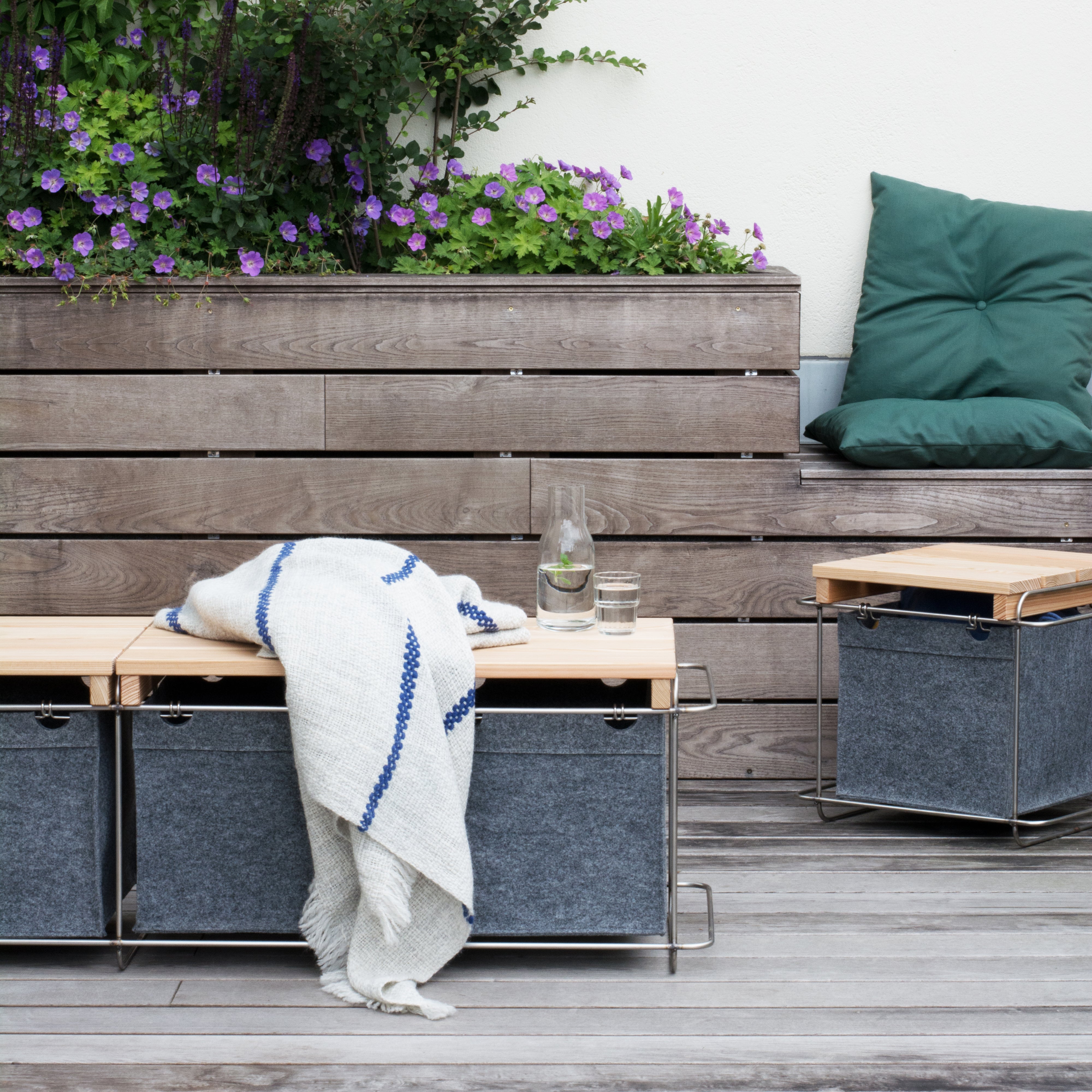 An outdoor patio setup features Bartmann Berlins Grit Stool Outdoor with modular wooden seating and a planter holding purple flowers. A glass pitcher, cushion, and blanket adorn the seating, while adjacent PET felt cushioned seats and wooden flooring complete the cozy scene.