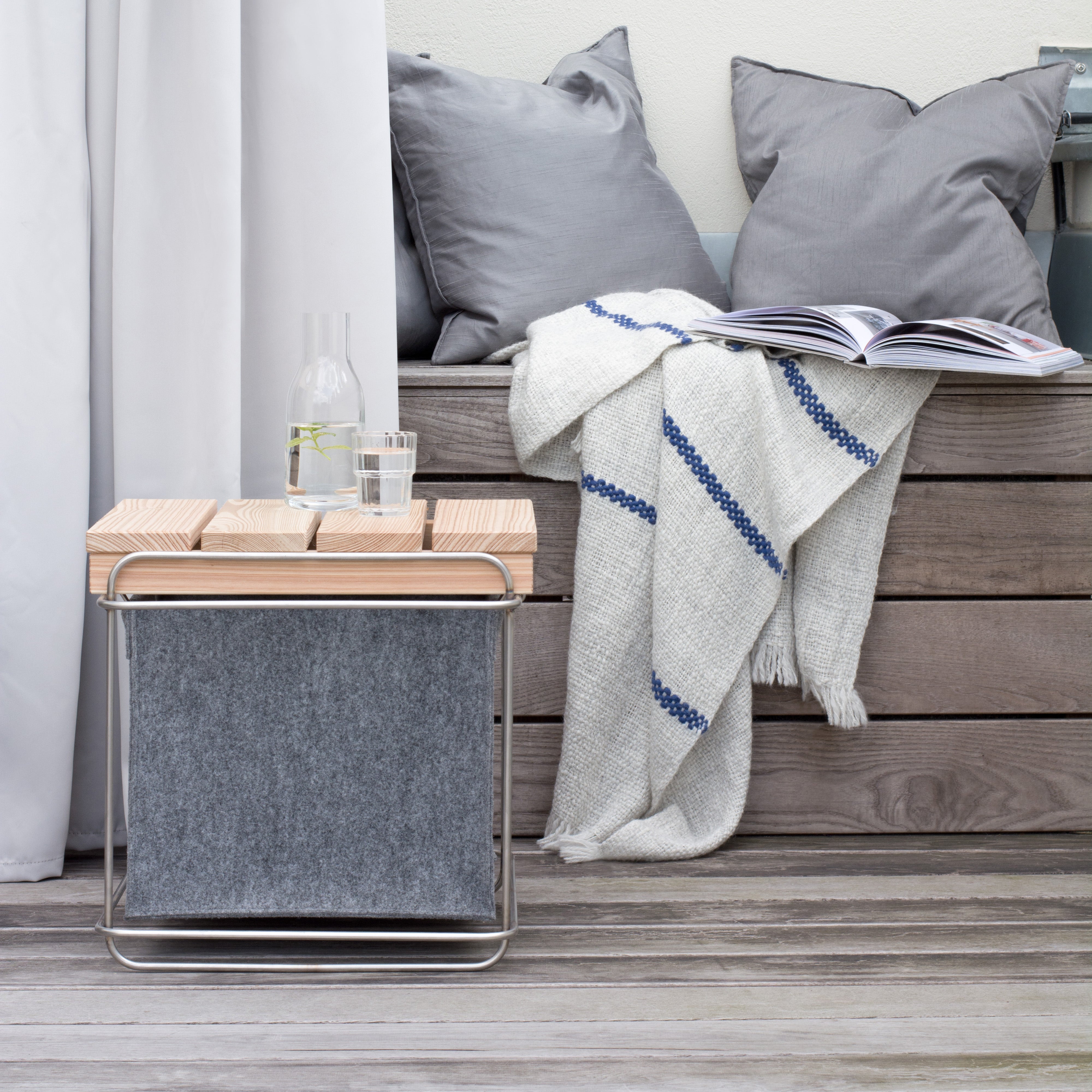 The Grit Outdoor collection by Bartmann Berlin transforms your outdoor space with gray cushioned benches and a striped blanket, complemented by a versatile side table holding a water carafe and glass. Enjoy customizable seating on elegant wooden decking, relaxing with an open book.