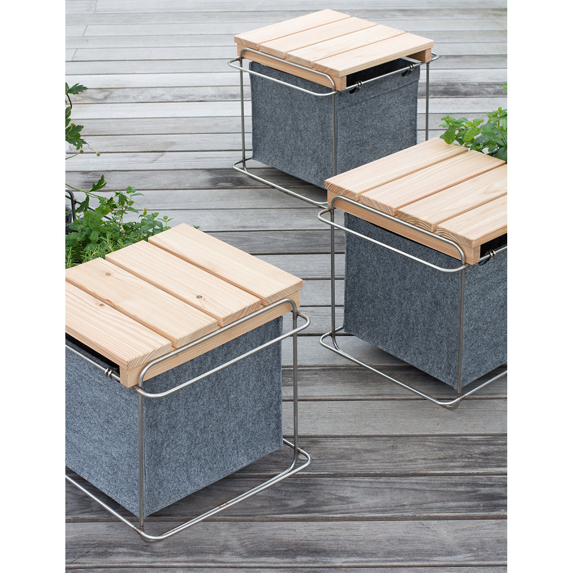Three Grit Outdoor planters by Bartmann Berlin, part of a versatile system with wooden tops and metal frames, feature gray fabric sides on a wooden deck. Small green plants peek from under the tops, enhancing the minimalist design.