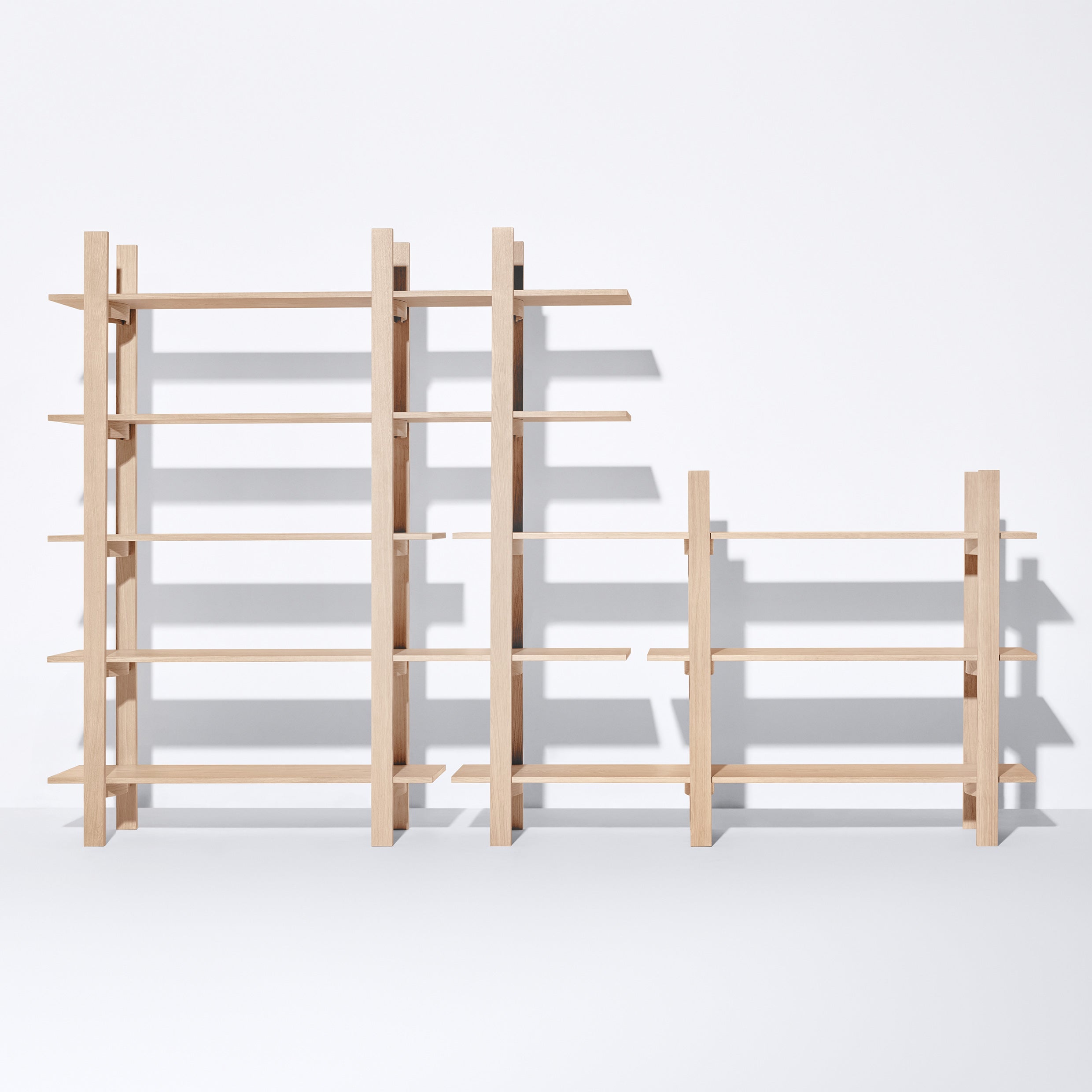 Freestanding Tonus wooden shelving units by Bartmann Berlin, varying in height, stand against a white backdrop. The minimalist design features sleek lines and solid oak open shelves, casting elegant shadows below.
