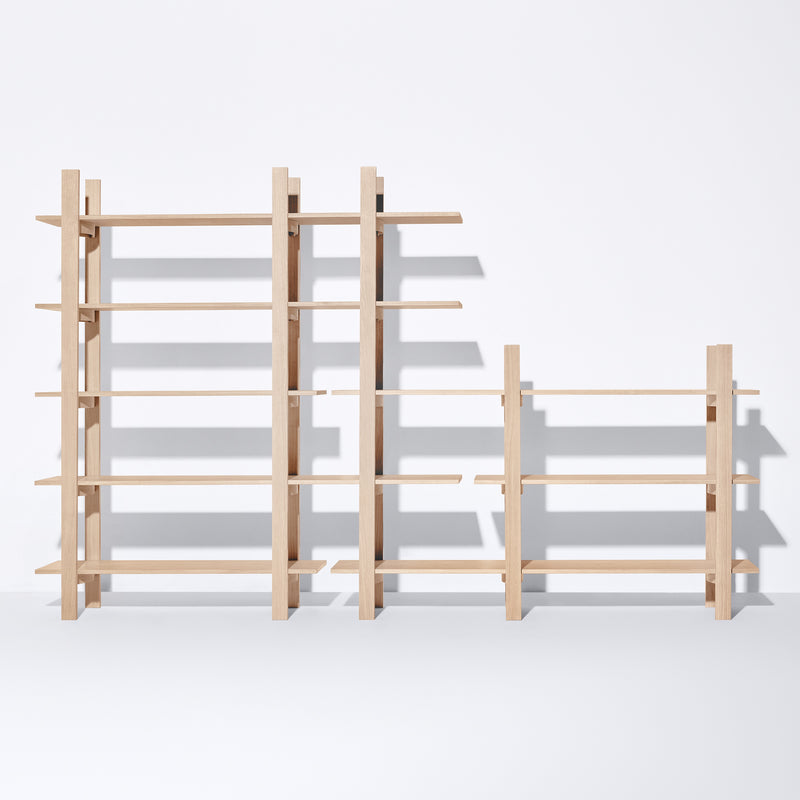 Freestanding Tonus wooden shelving units by Bartmann Berlin, varying in height, stand against a white backdrop. The minimalist design features sleek lines and solid oak open shelves, casting elegant shadows below.