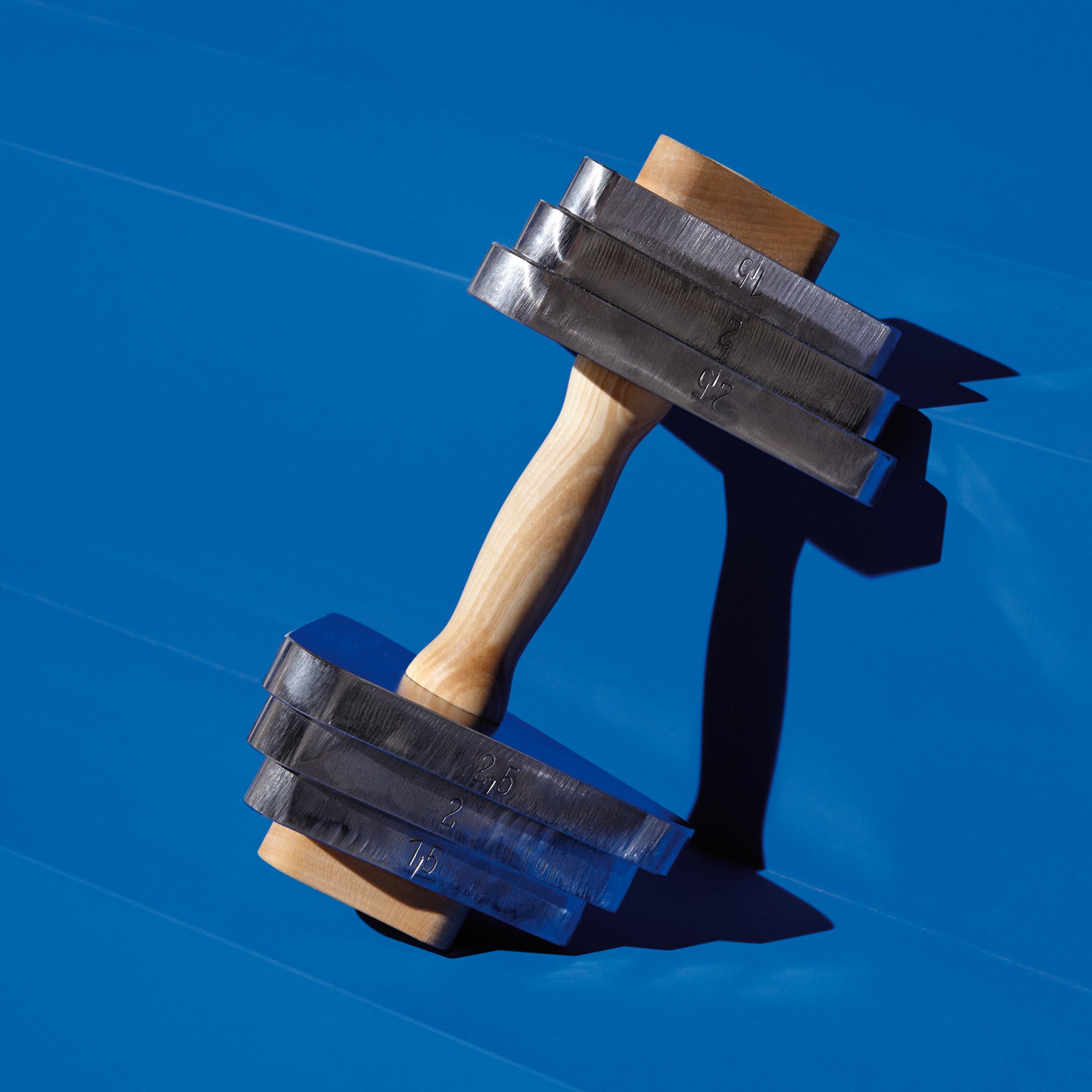 A Bartmann Berlin Trimm Dich dumbbell with a maple handle and four rectangular metal plates on each side sits on a bright blue surface, with numbered weights casting shadows.