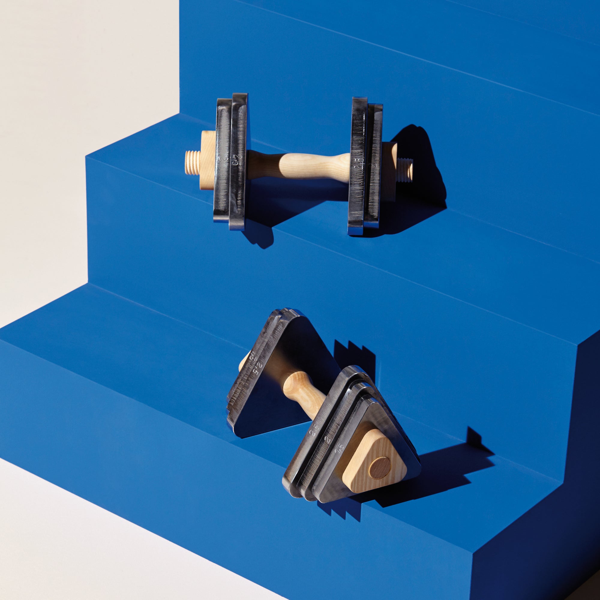 Two triangular Trimm Dich barbells by Bartmann Berlin, with maple wood handles, rest on a blue, stair-like structure. The lighting casts defined shadows, highlighting the contrast between the barbells and the bright blue background.