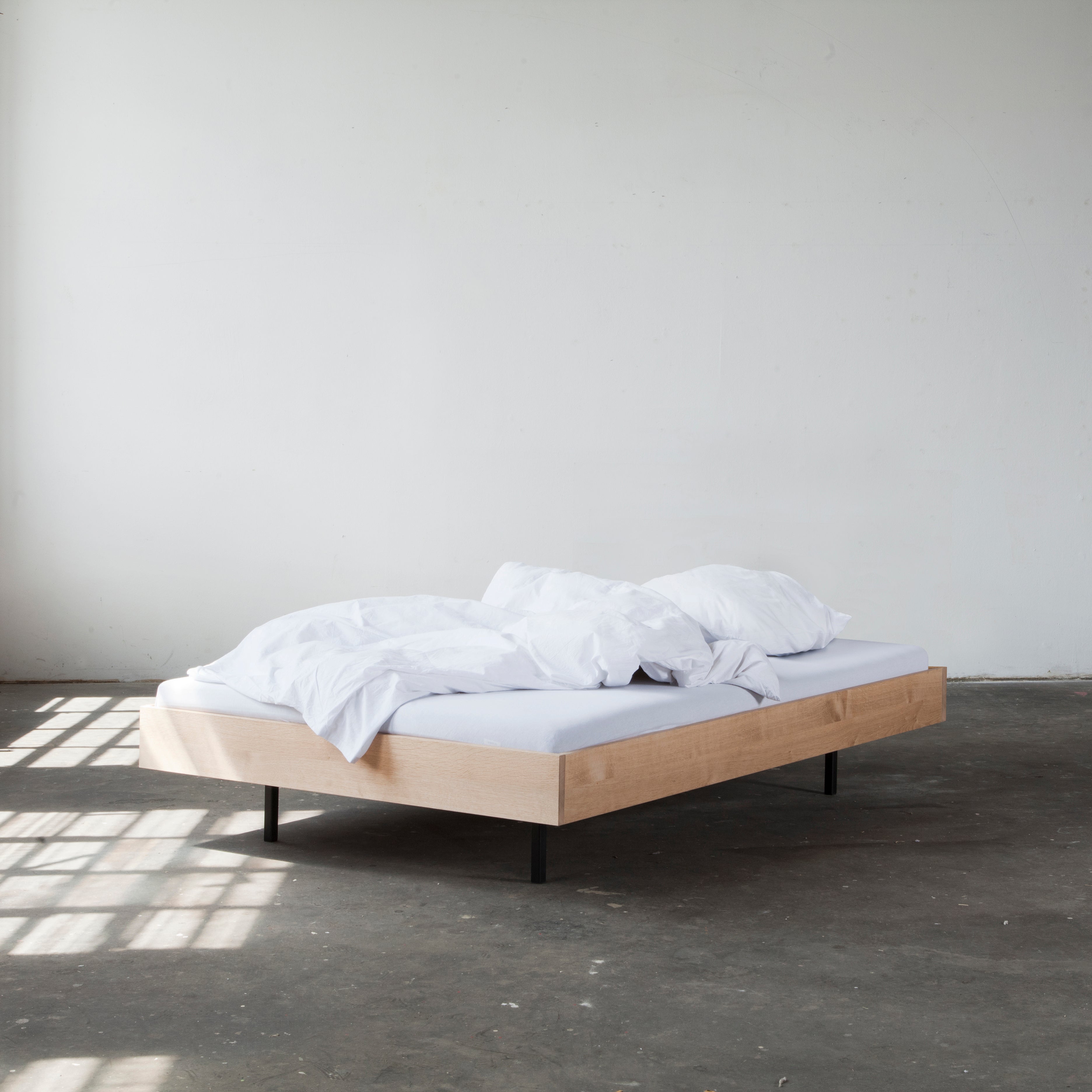 A minimalist bedroom scene showcases the Bartmann Berlin Unidorm Bed without Headboard, made of oiled oak with white bedding and pillows against an off-white wall. Sunlight streams through a window, casting shadows that emphasize its elegant design.