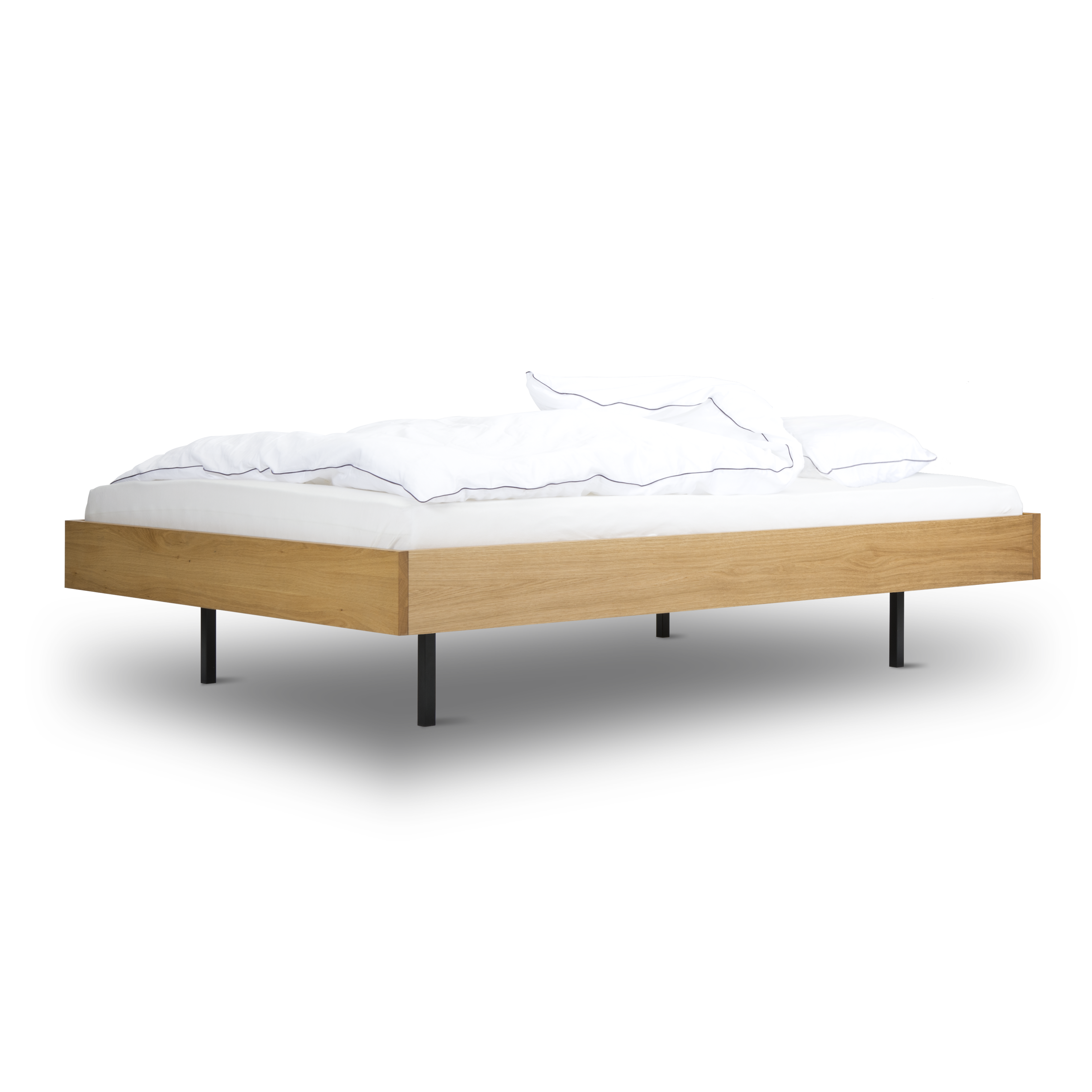 The Unidorm Bed without Headboard from Bartmann Berlin features a modern wooden frame made of oiled oak with powder-coated steel legs, paired with a white mattress and crumpled white bedding, all set against a transparent background.