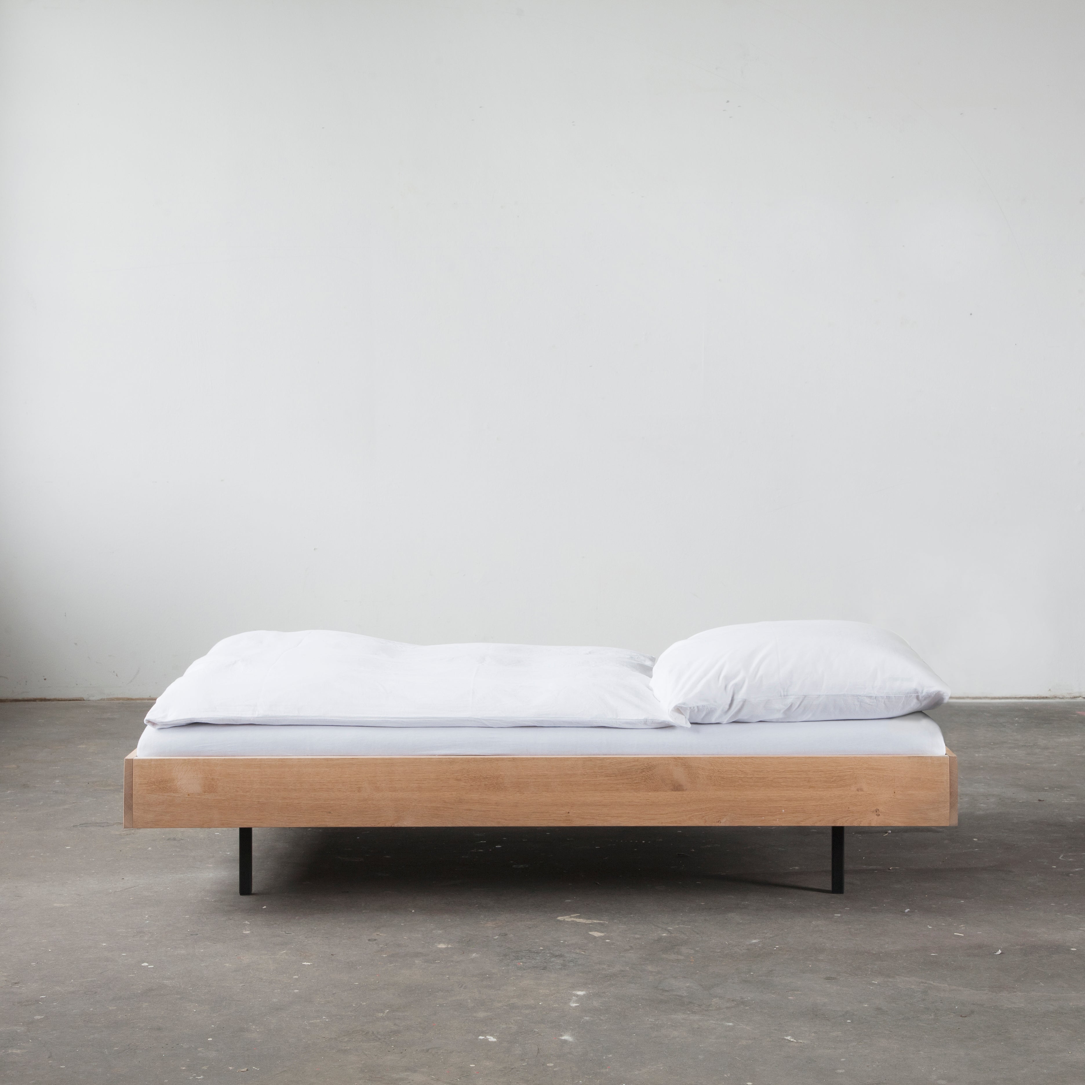 The Unidorm Bed without Headboard by Bartmann Berlin features a minimalist wooden frame made of oiled oak, complemented by powder-coated steel legs. This low-profile bed includes a white mattress, pillow, and duvet, set on a plain concrete floor against a white wall for a simple, modern look.