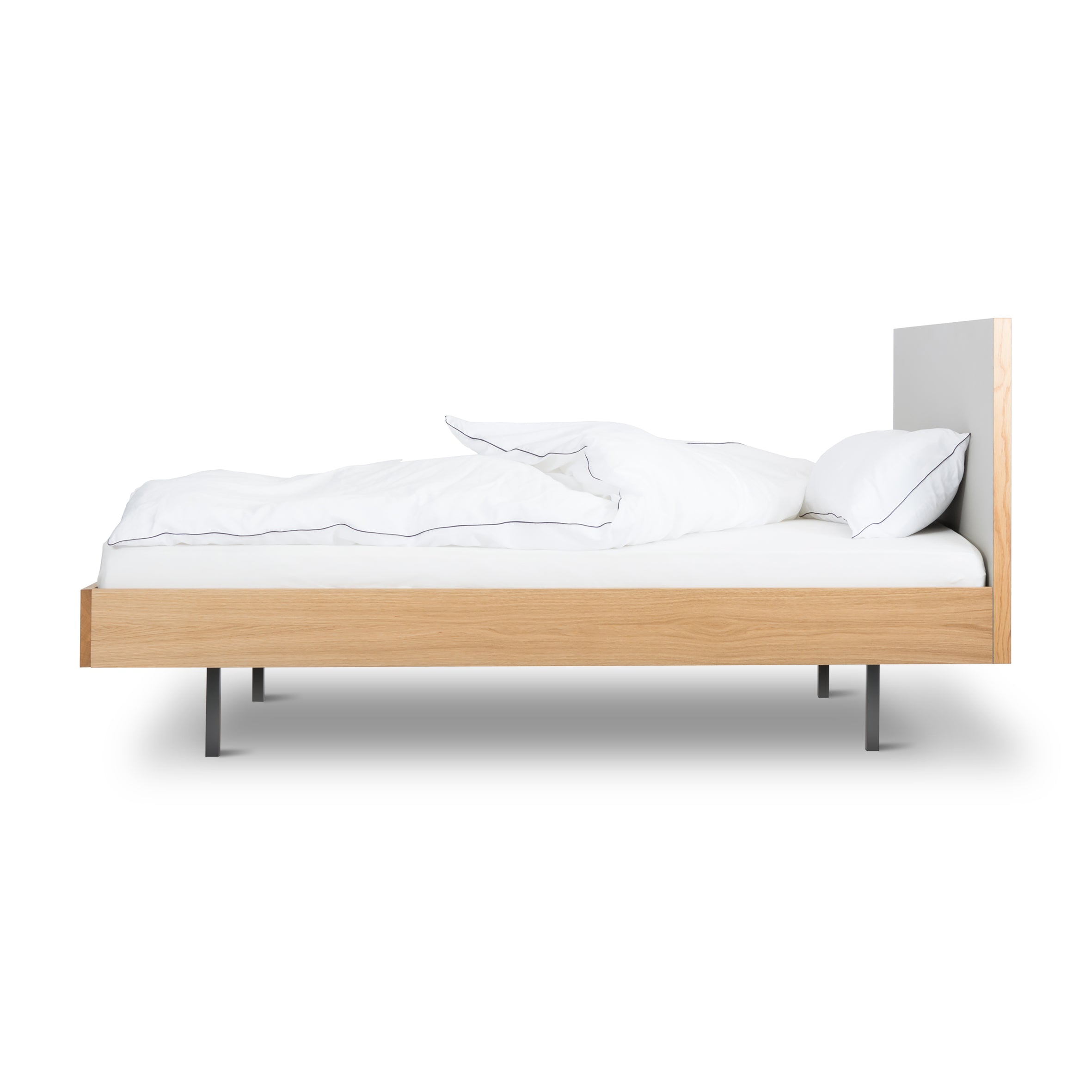 The minimalist Unidorm Bed with Headboard by Bartmann Berlin, crafted from oiled oak with sleek powder-coated steel legs, showcases neatly arranged white bedding and pillows. Photographed from the side against a plain white background, it embodies elegant simplicity.