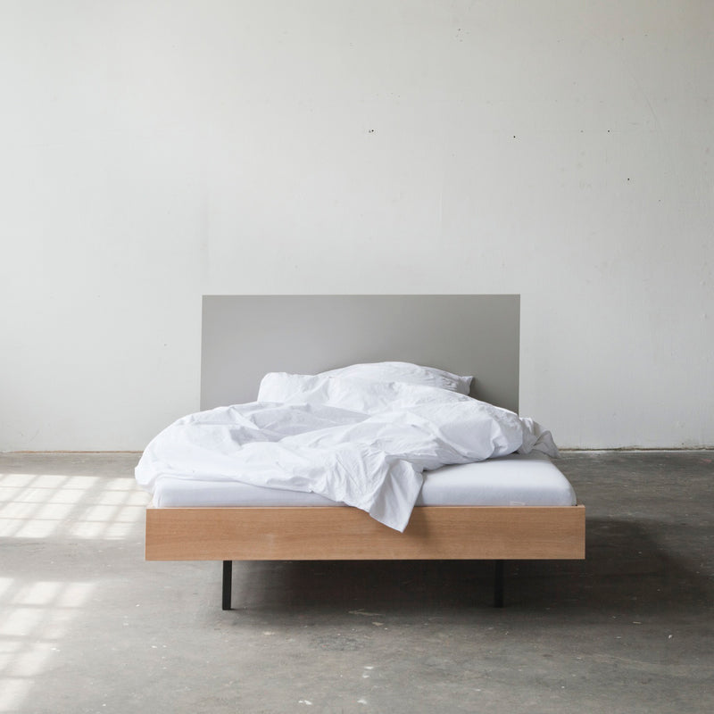 In a minimalist bedroom, the Unidorm Bed with Headboard from Bartmann Berlin showcases an unmade bed with white linens on a light wooden frame made of oiled oak, against a bare concrete floor. Sunlight streams through a window, casting shadows and heightening the rooms simple elegance.