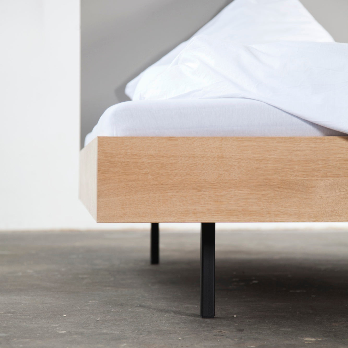 Set against a white and gray backdrop, the Bartmann Berlins Unidorm Bed with Headboard, featuring oiled oak and black powder-coated steel legs, is adorned with white bedding and mattress, resting elegantly on a smooth concrete floor.