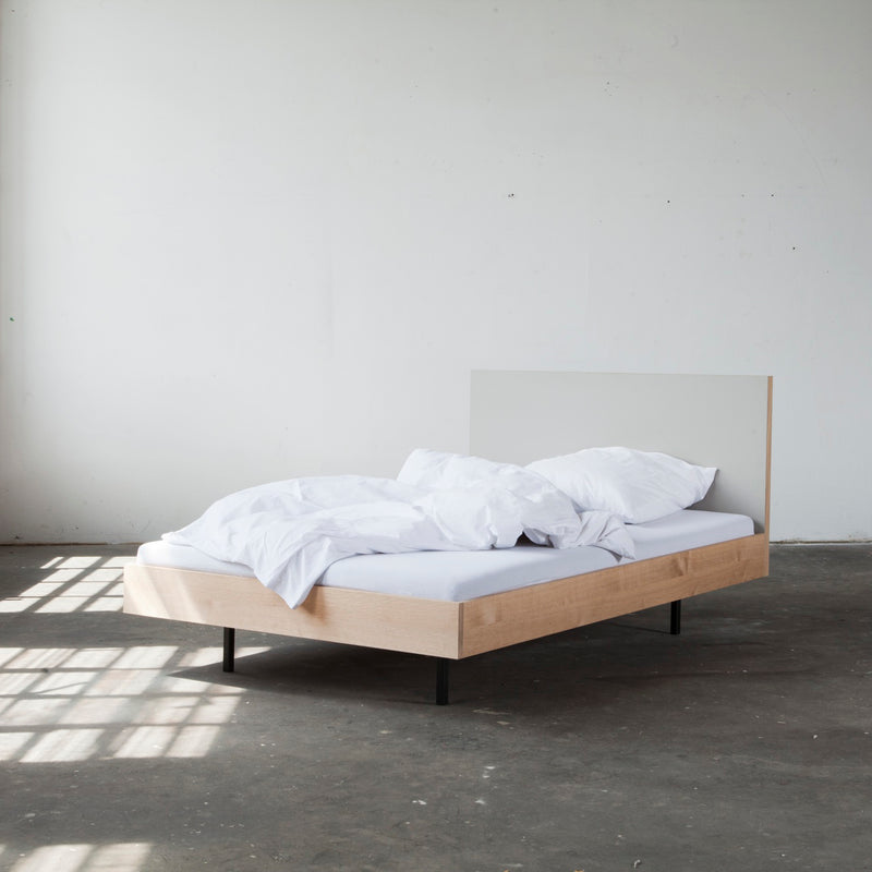 In a minimalist room, the Bartmann Berlin Unidorm Bed with Headboard in oiled oak is complemented by white sheets, pillows, and a comforter—all slightly rumpled. Sunlight streams in, casting shadows on the concrete floor. The plain white walls complete the serene retreat.