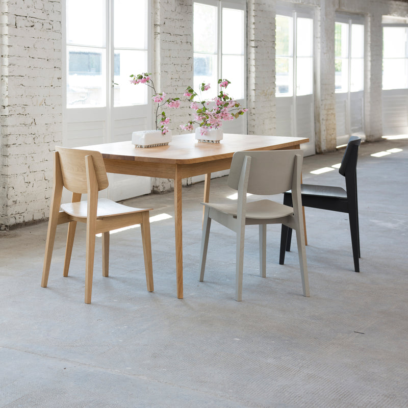 A wooden table exudes natural elegance with three Usus chairs in light wood, gray, and black hues. The bright room, featuring white brick walls and large windows, is adorned with two vases of pink flowers, capturing Bartmann Berlins refined style.